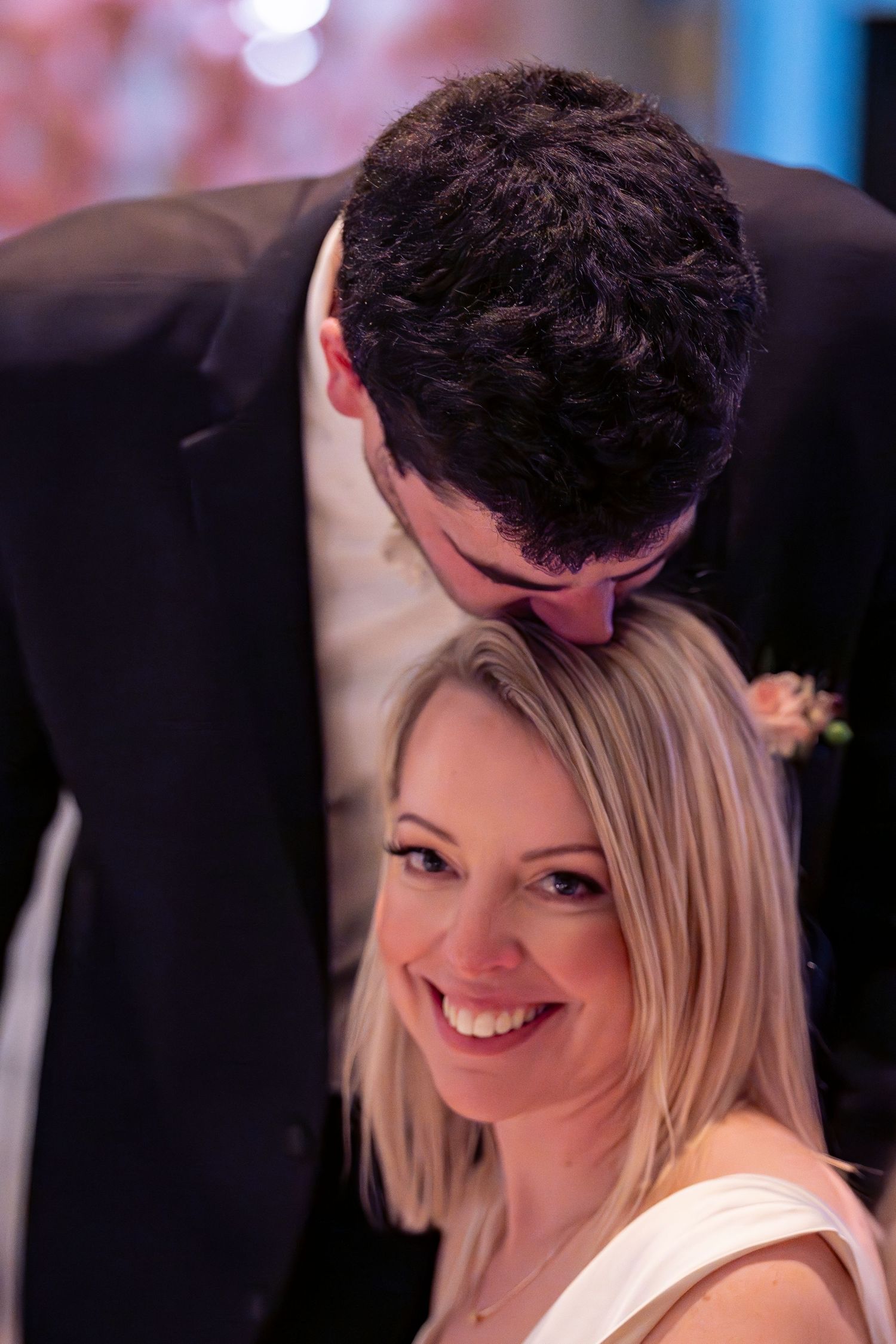 Groom tenderly kisses bride on the head as she smiles warmly during their wedding reception.