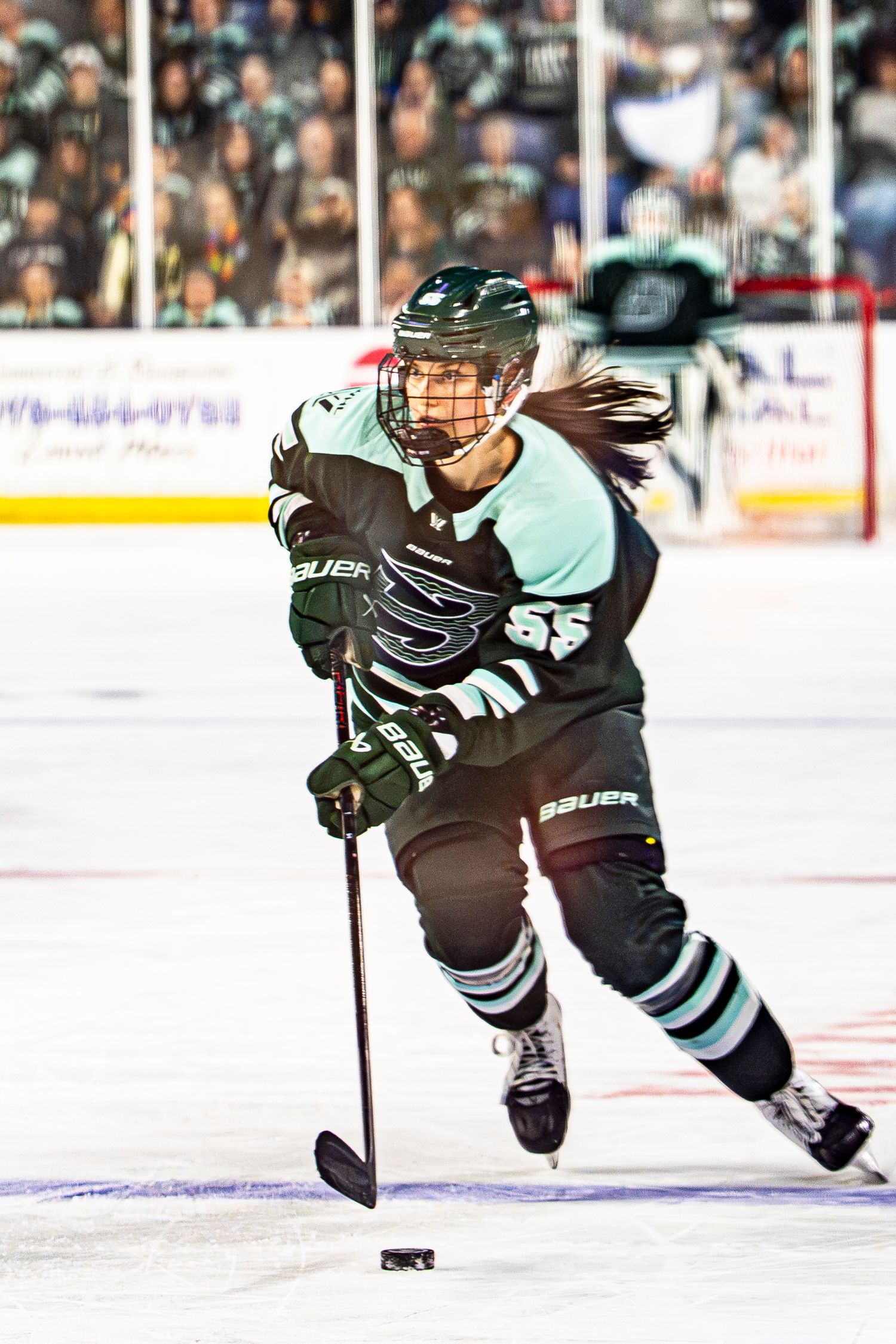 Female hockey player in green Bauer gear skates with the puck during an indoor game.