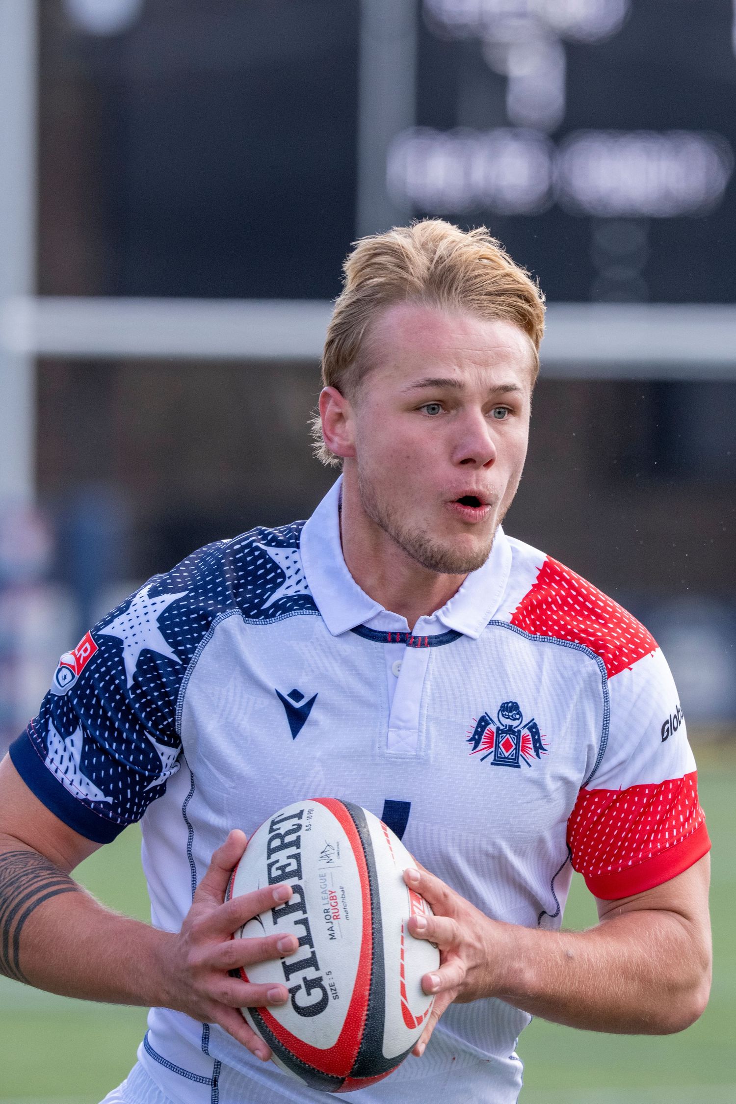 Blonde rugby player in stars and stripes jersey carrying ball during outdoor match.