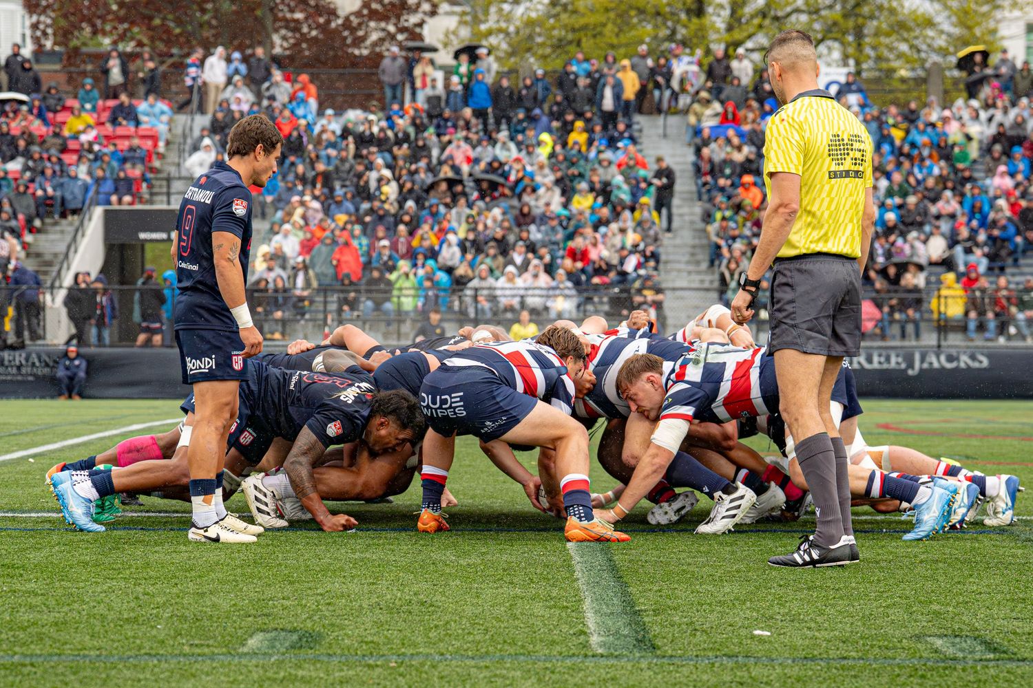 Rugby players engage in a scrum during a competitive match before a large crowd of spectators at an outdoor stadium.
