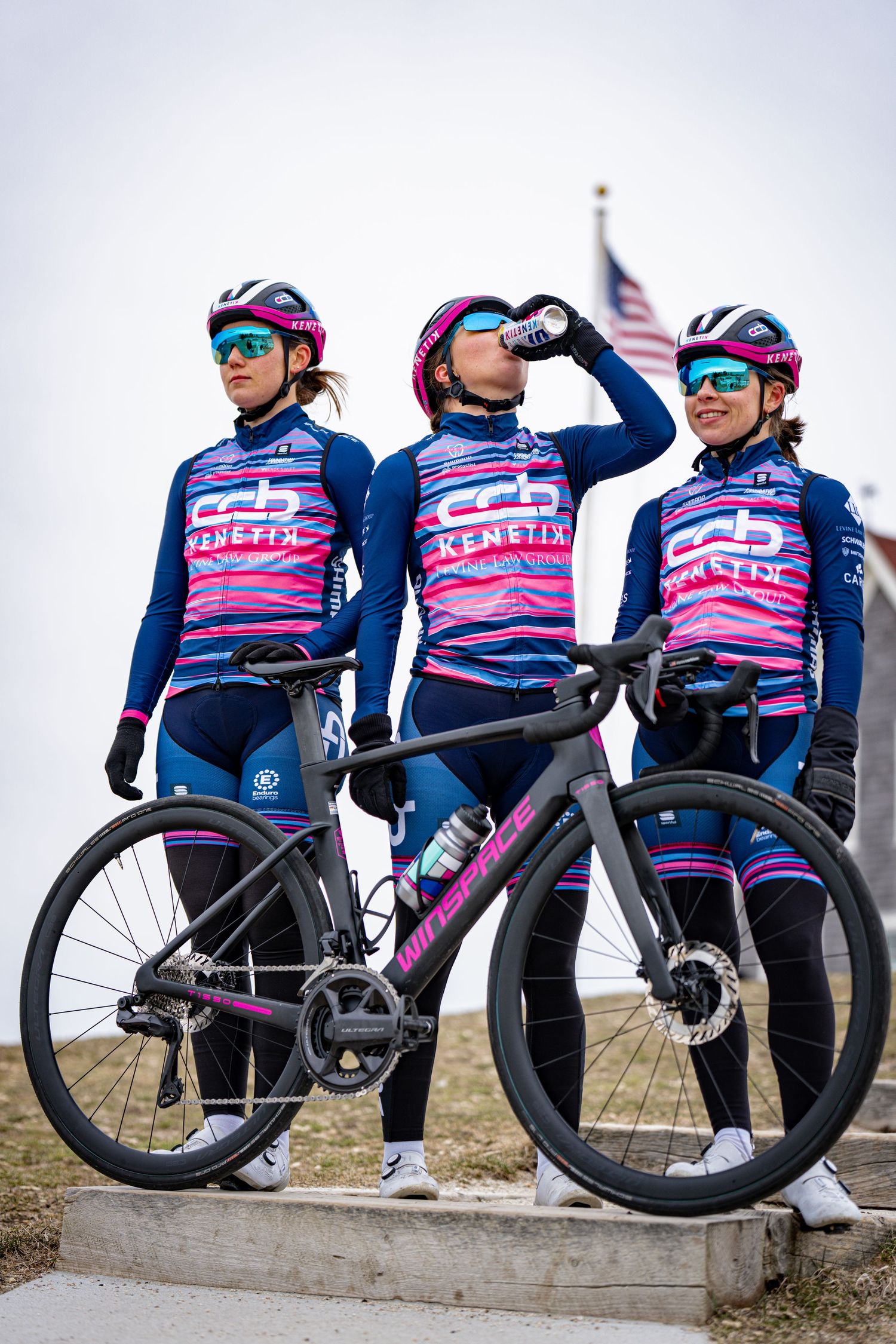 Three female cyclists in pink and blue CB Genetix team kits posing with road bike outdoors.