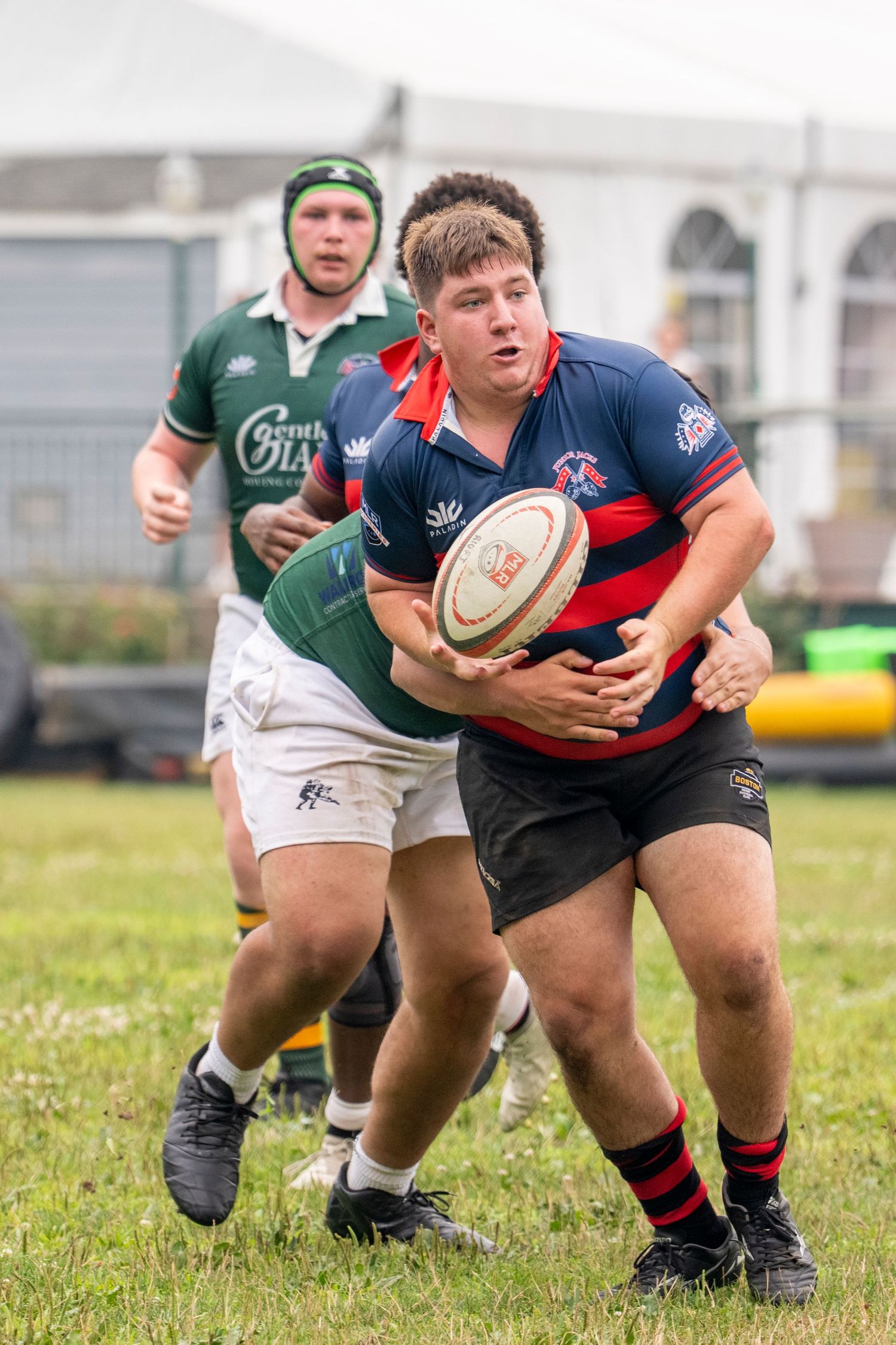 Rugby player in navy and red jersey carries ball while being tackled during an outdoor match.