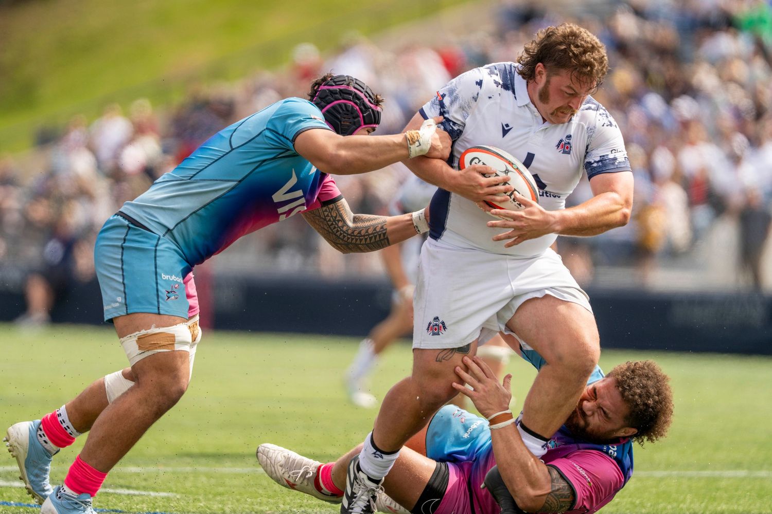 Rugby player in white carries the ball while being tackled by two opponents.