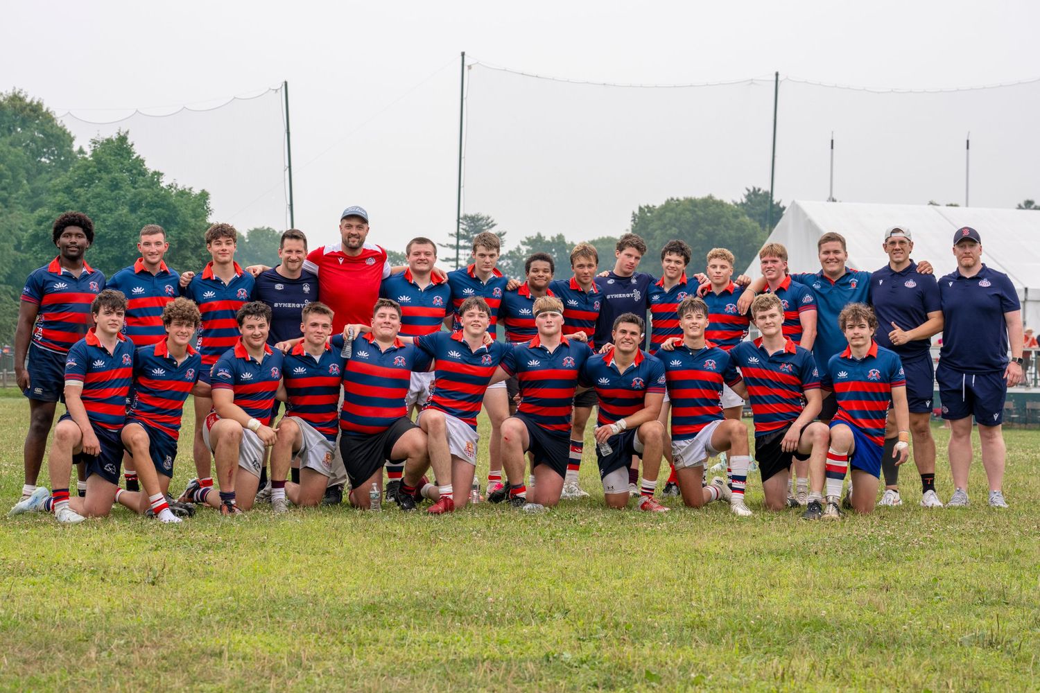 Youth rugby team poses together on a grass field wearing red and blue striped jerseys with coaches.
