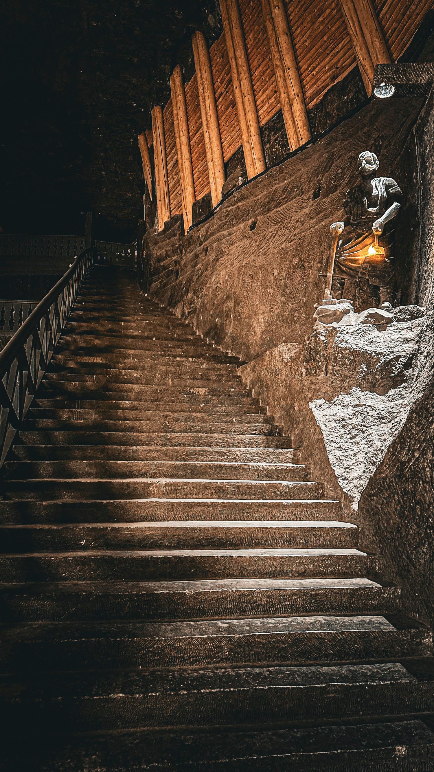 A dramatic staircase in Wieliczka Salt Mine with a miner statue holding a lantern beside carved stone walls.