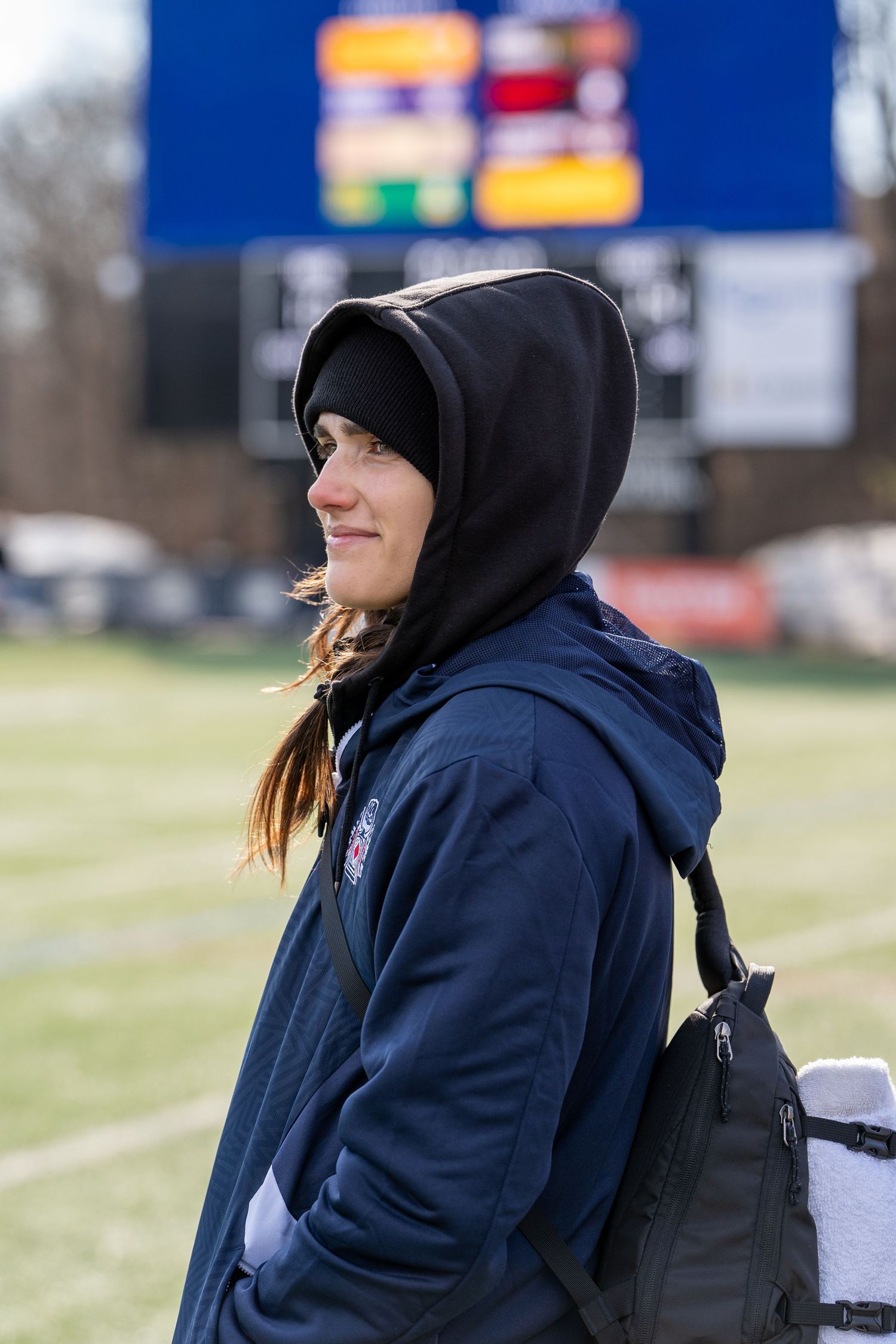 Young woman in navy hoodie with backpack standing on sports field near scoreboard.