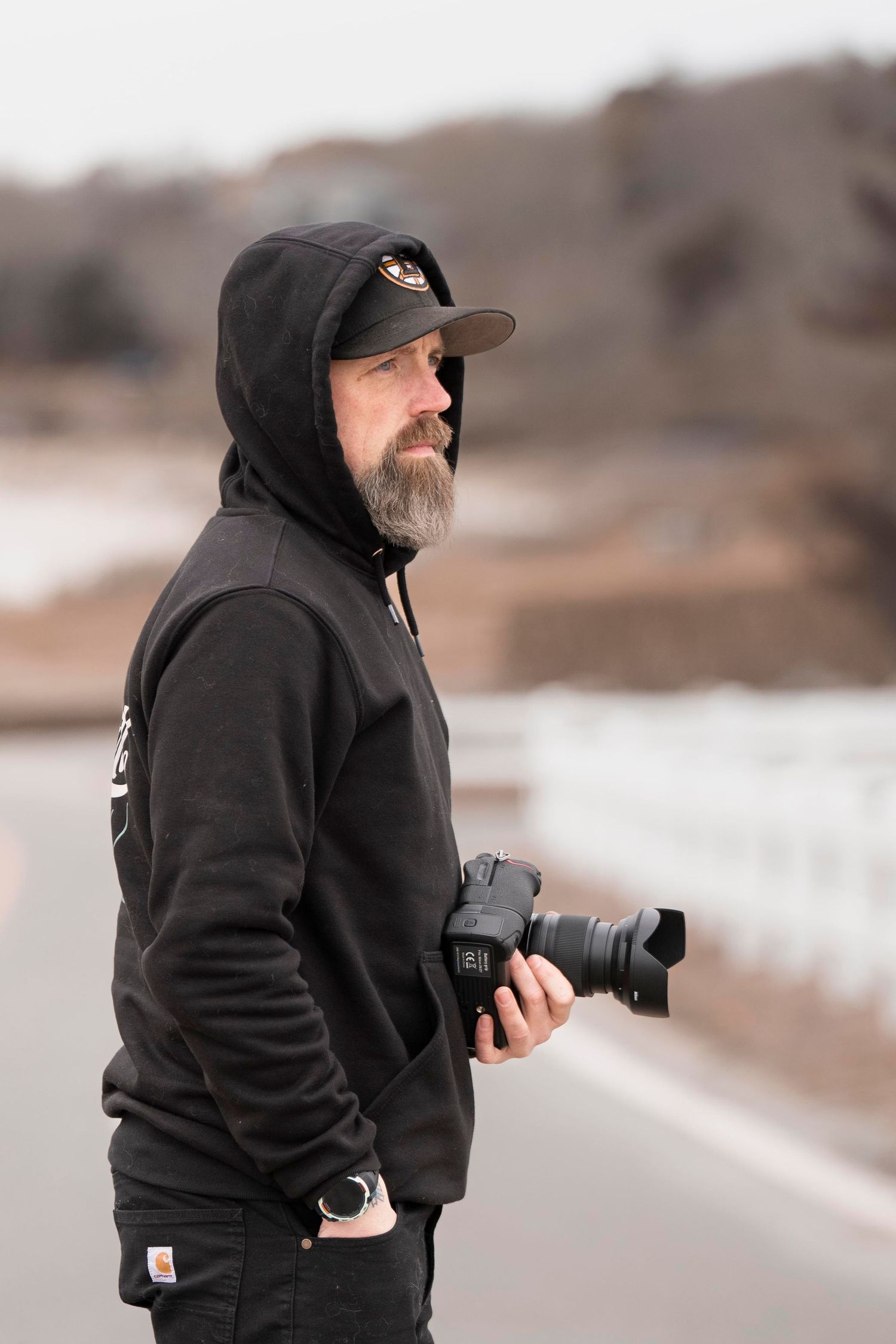 Bearded man in black hoodie and cap holds DSLR camera outdoors on overcast day, standing on road with blurred winter background.