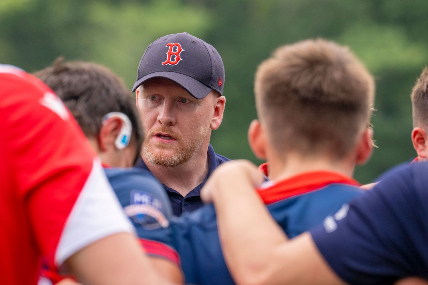 A coach in a Boston Red Sox cap addresses rugby players during a team huddle outdoors.