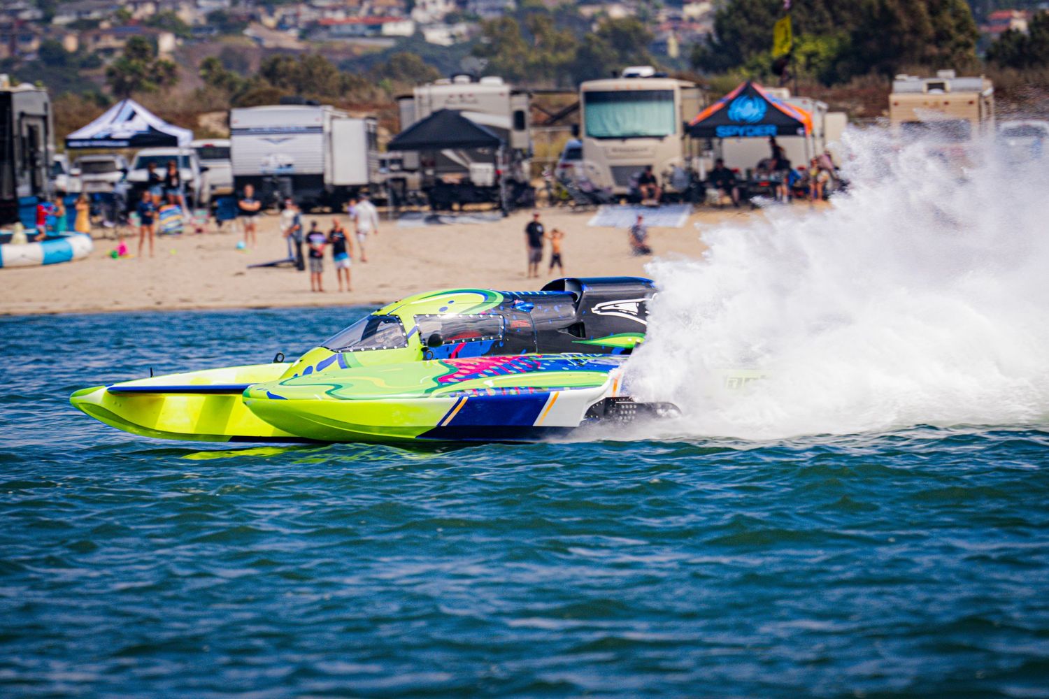 Bright yellow-green racing powerboat competes at high speed near a sandy beach during a race.