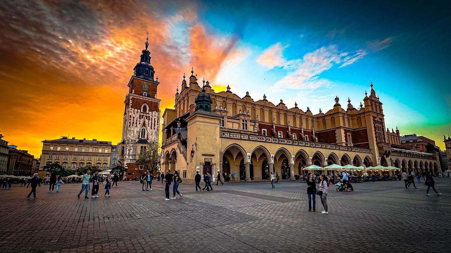 Krakow Main Market Square at sunset with colorful sky, historic Cloth Hall and Town Hall Tower surrounded by tourists.