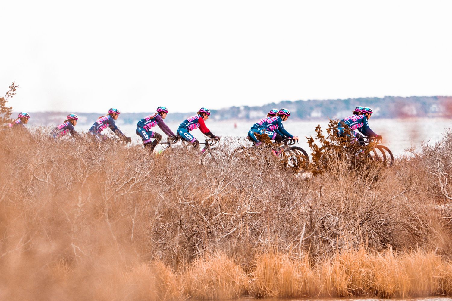 Cyclists race through a dry desert landscape with sparse brown shrubs and a lake visible in the background.