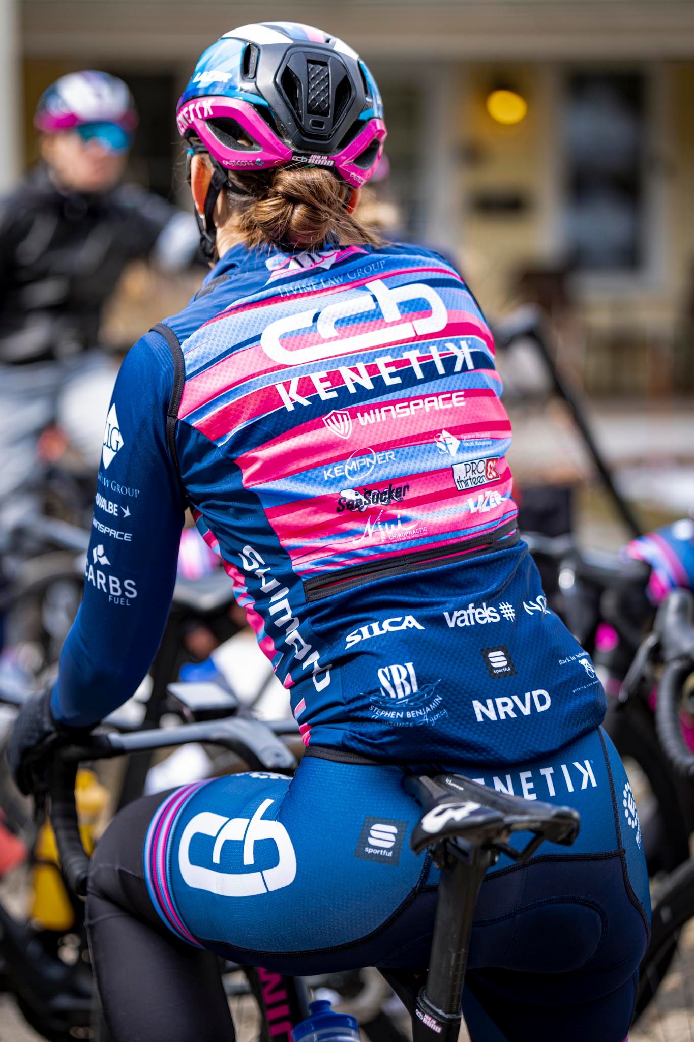 Female cyclist wearing CCB Kenetik team jersey in blue and pink, sitting on bike at cycling event.