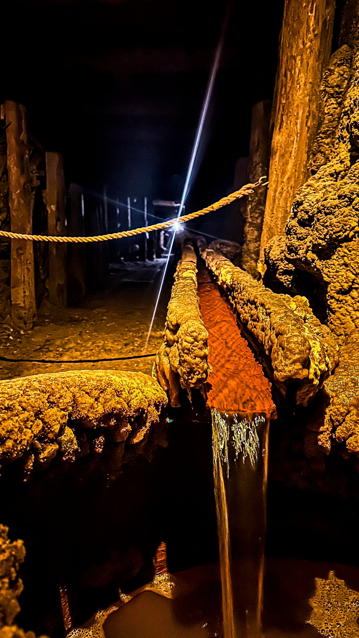 Underground cave with mineral-encrusted wooden beams and flowing water illuminated by a flashlight.