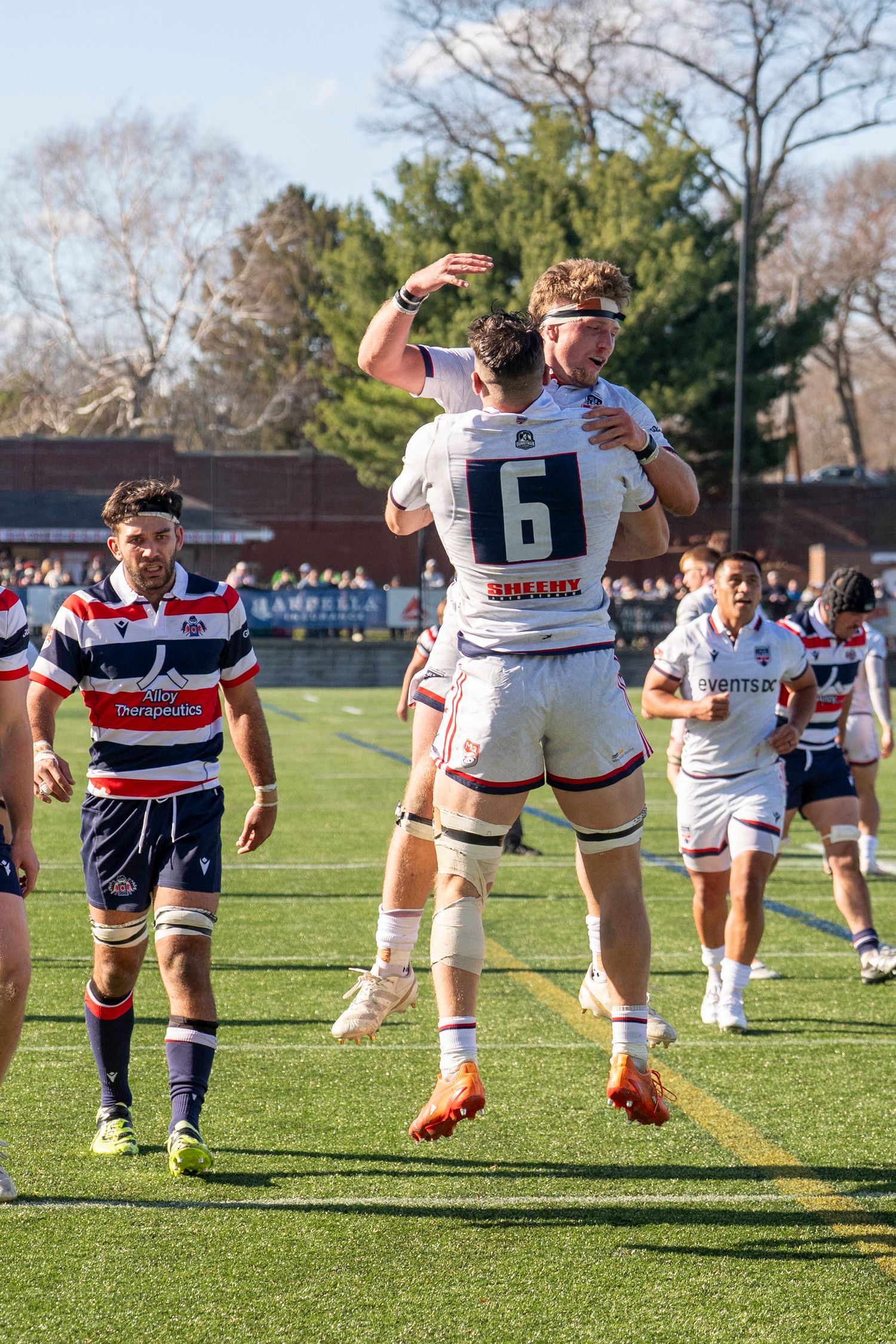 Rugby players in white jerseys celebrate with a jump hug during a match on a green field.
