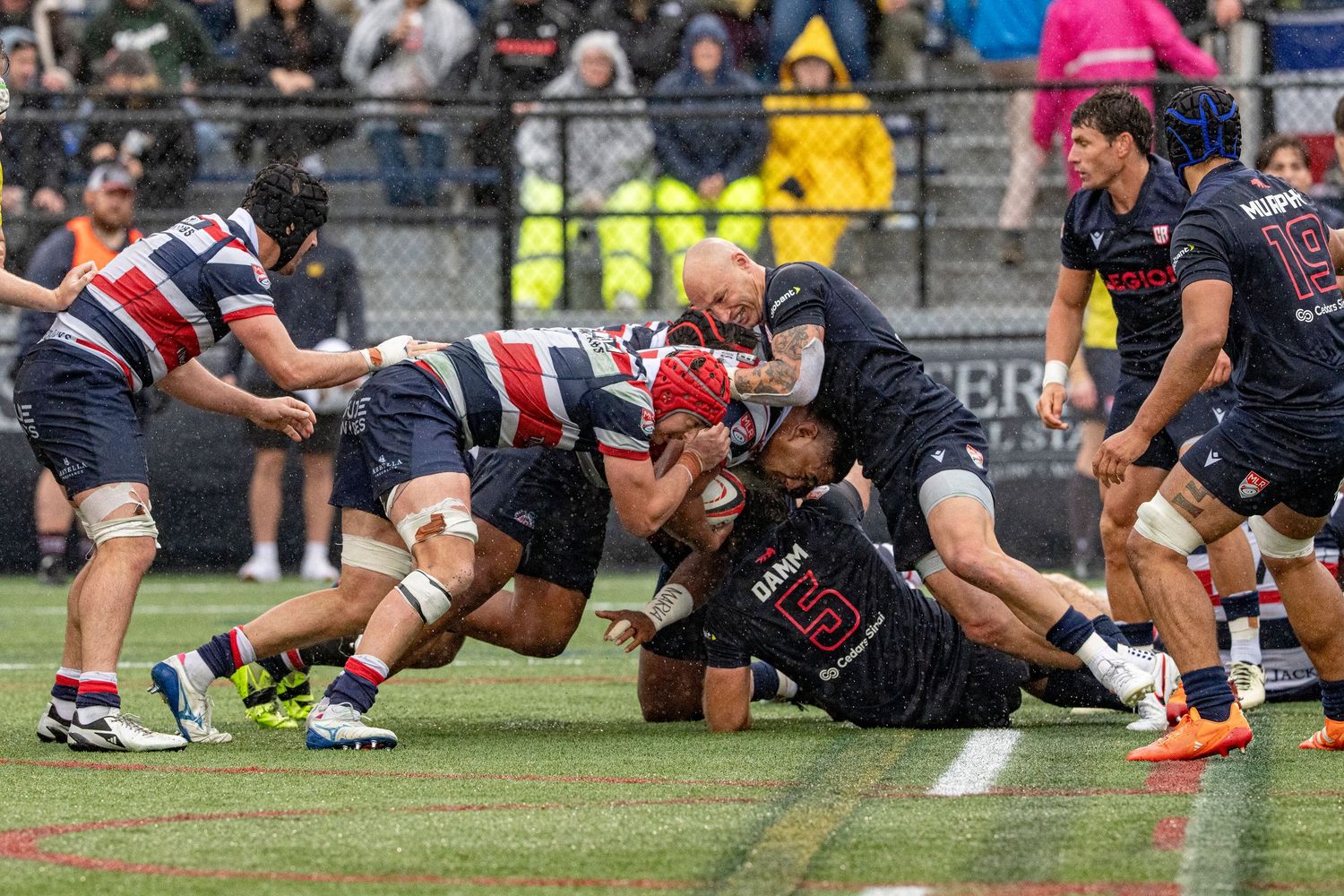 Rugby players engage in an intense tackle and maul during a competitive match in wet conditions.