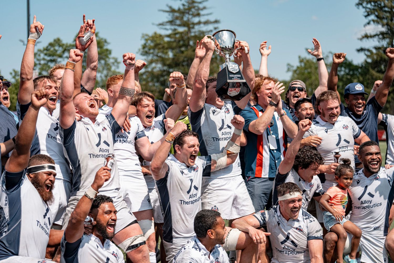 Rugby team celebrates a championship victory, lifting a trophy while cheering in sunny weather.