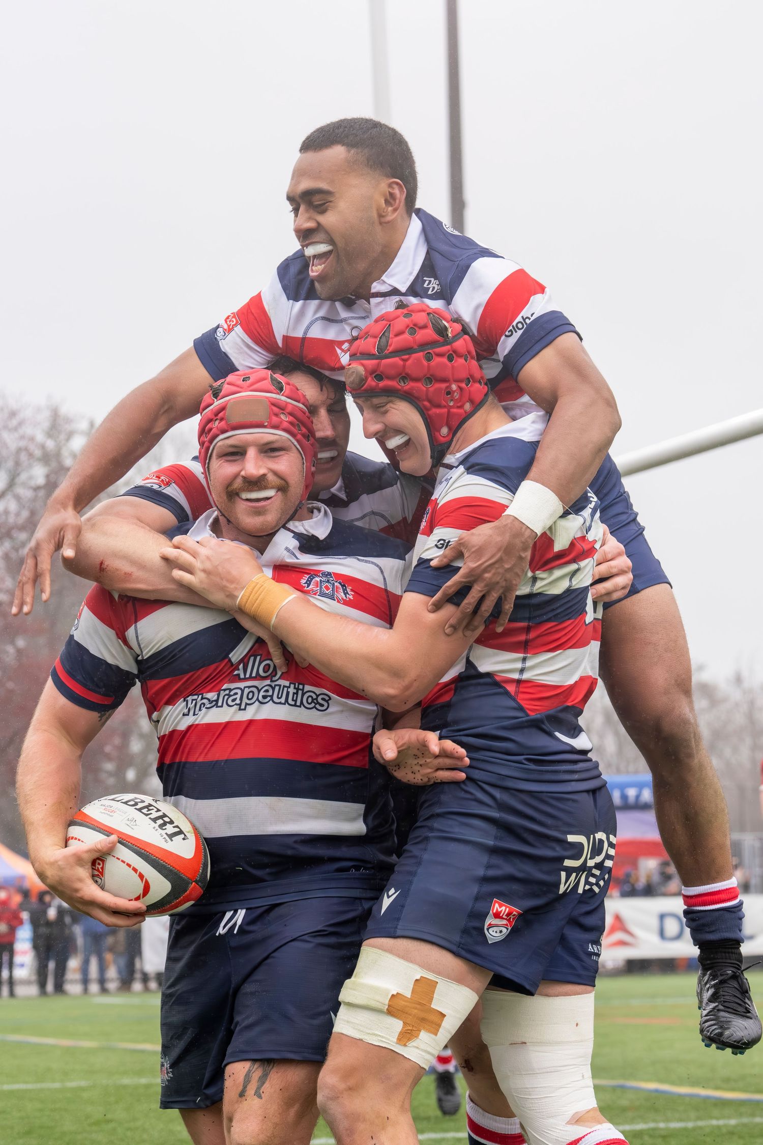 Rugby players celebrate try score in rainy match, hugging joyfully in red and blue uniforms.