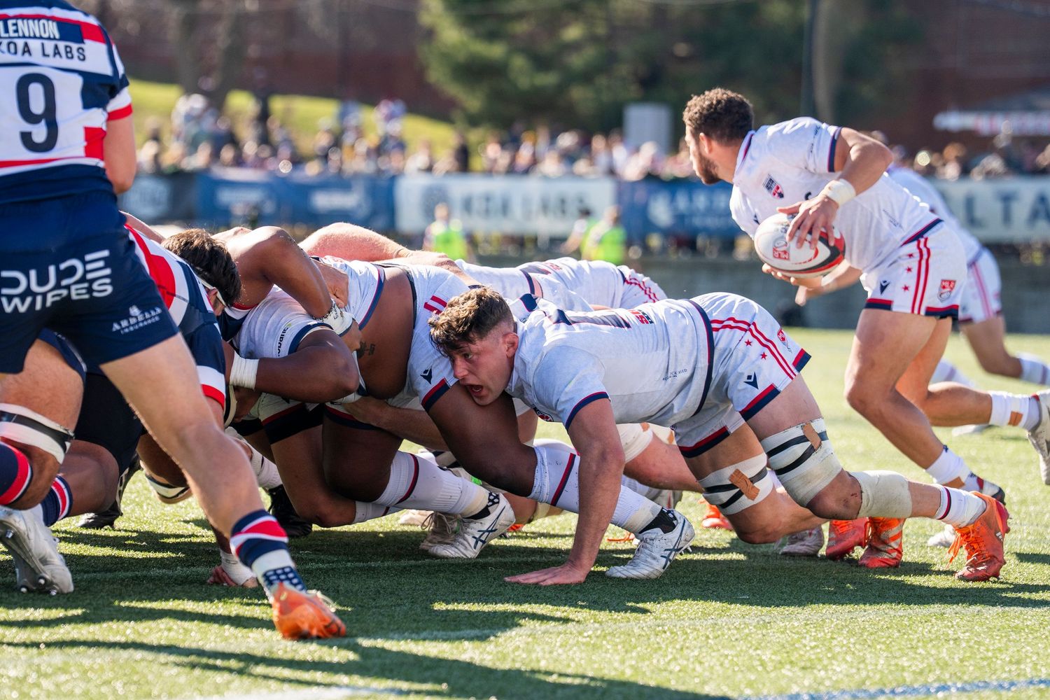 Rugby players compete in an intense scrum during a match, with spectators watching from the sidelines.