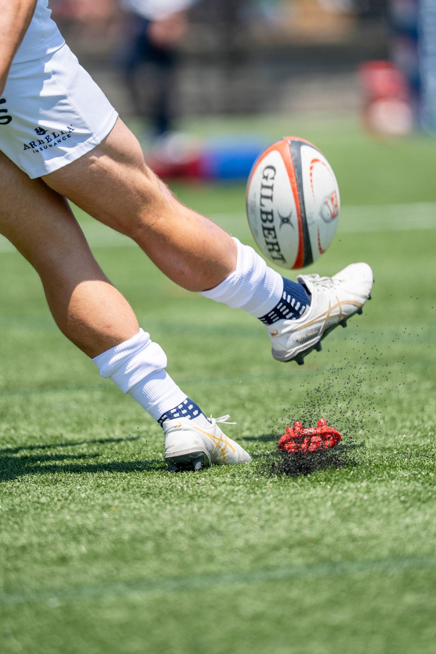 Rugby player kicking a Gilbert ball off a tee on a green field during a match.