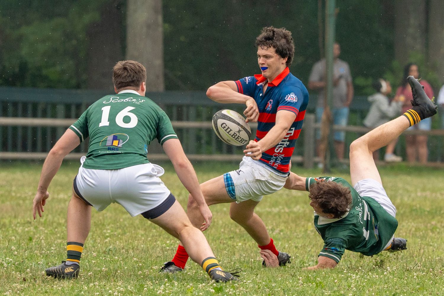Rugby players in green and red uniforms compete for the ball during an outdoor match on a grass field.