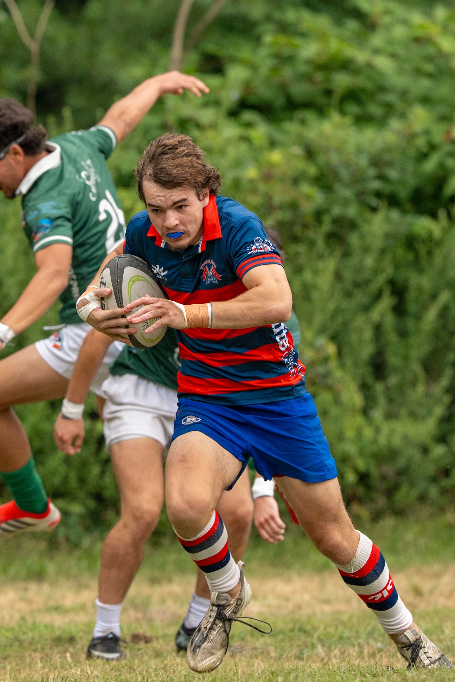 Rugby player in blue and red jersey carries ball while evading green-uniformed opponents during an outdoor match.