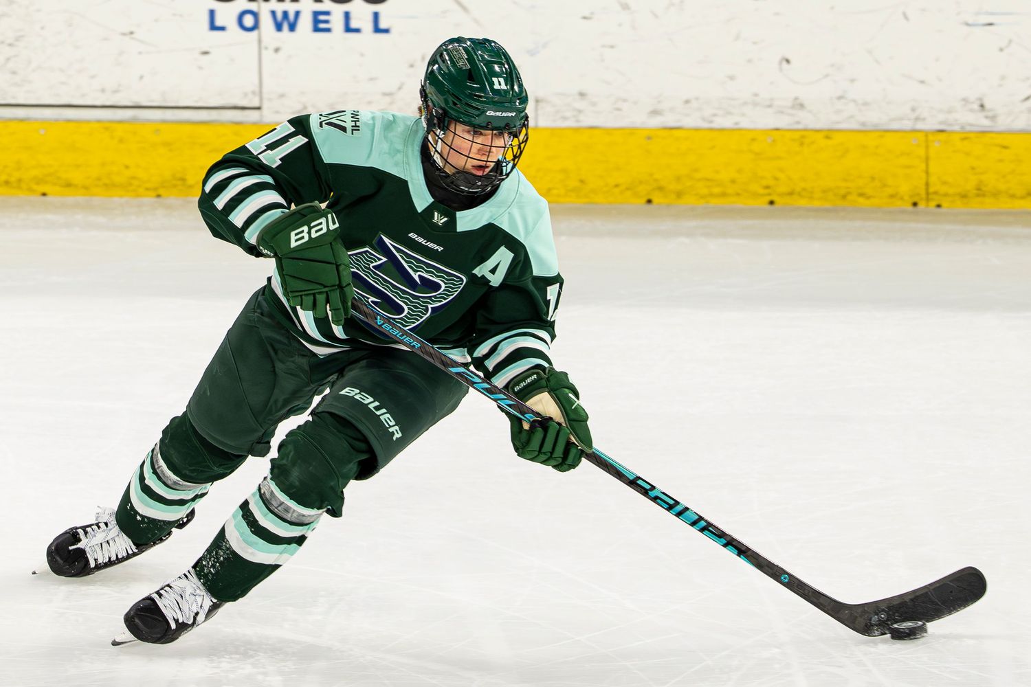 Female hockey player in green uniform skating with puck during a competitive ice hockey game.