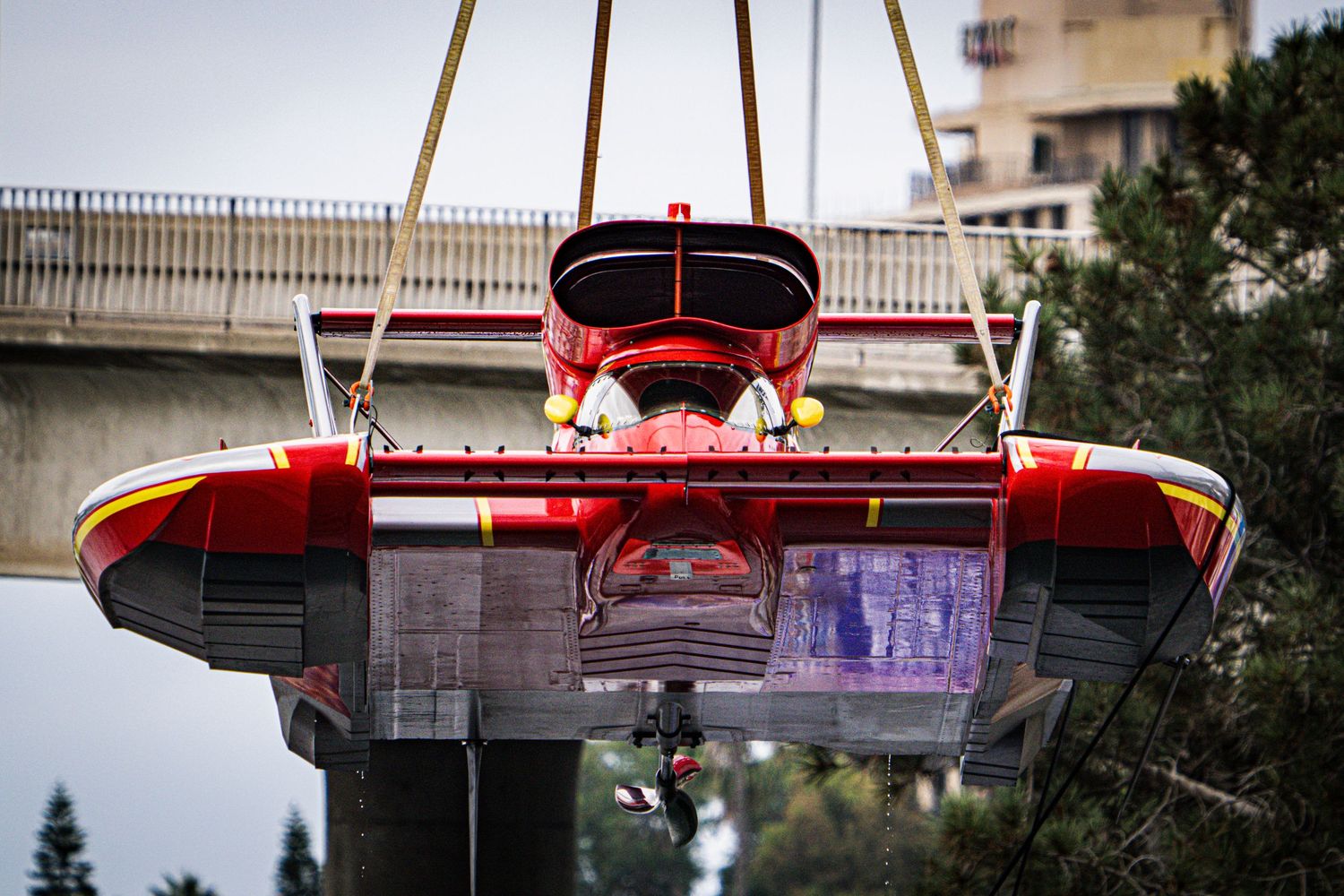 Red firefighting aircraft suspended by crane beneath highway overpass during operation.