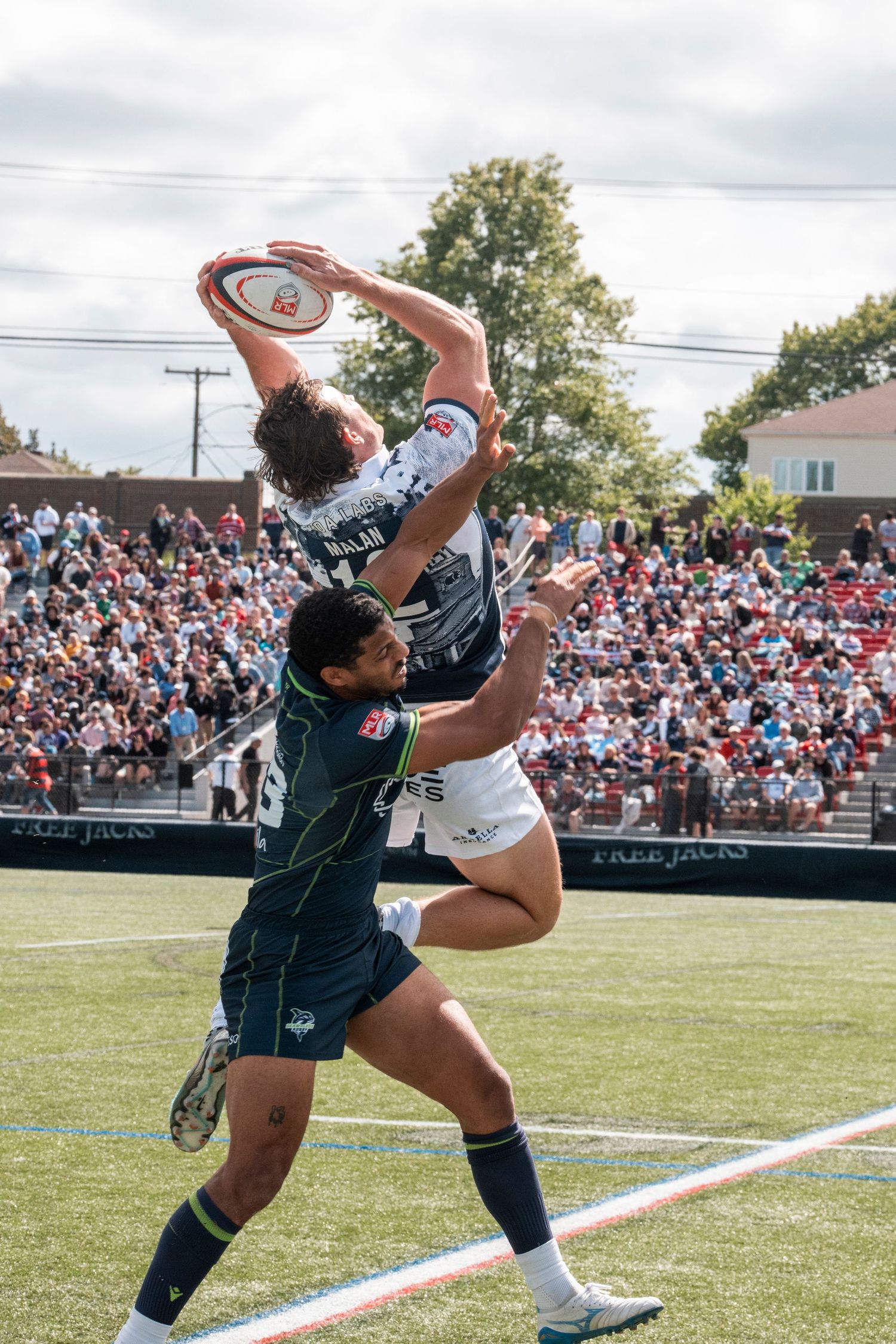 Rugby players compete for high ball catch during packed stadium match with Free Jacks team.