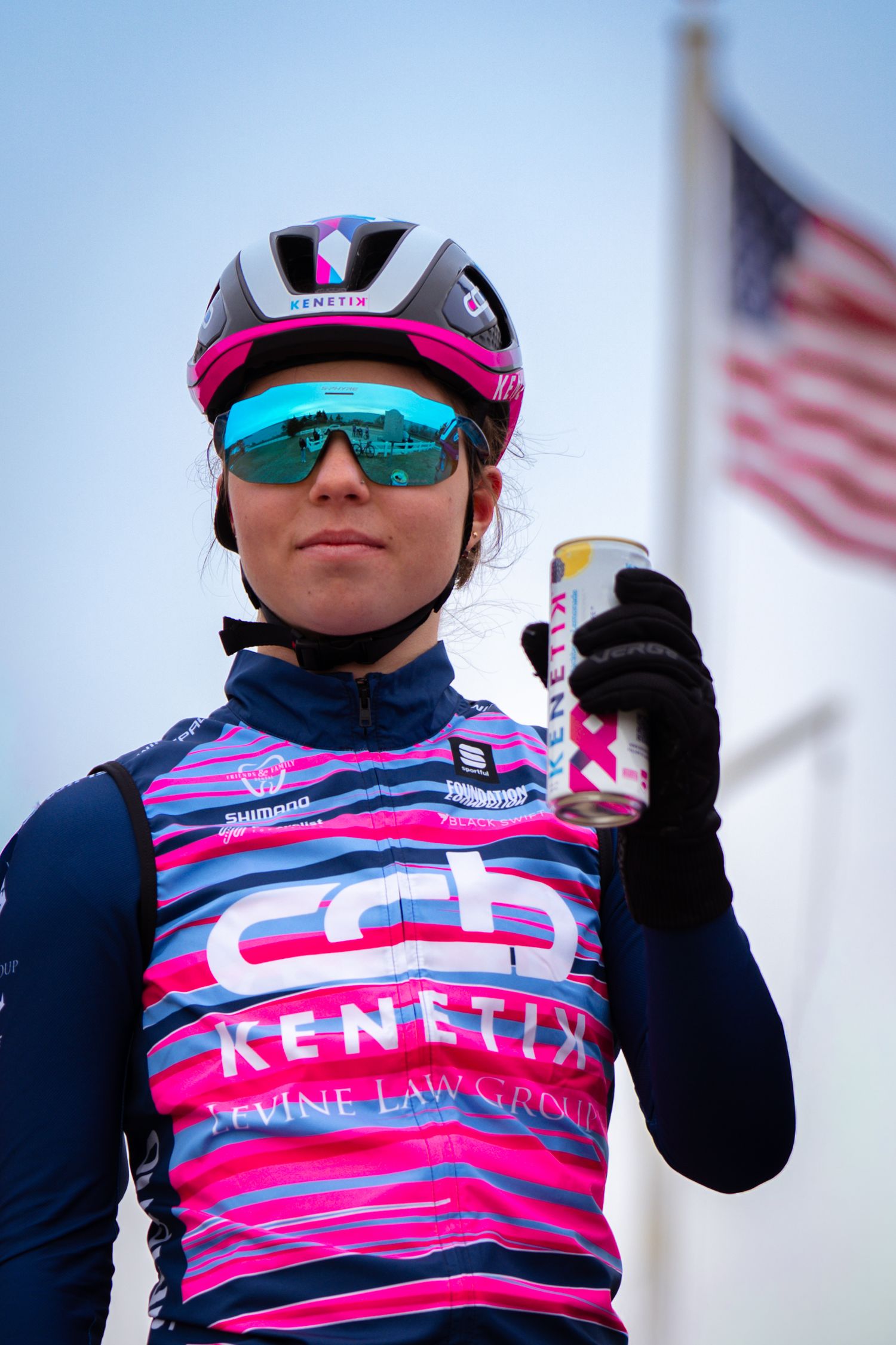 Female cyclist in CCB Kenetix team kit holding energy drink with American flag background.