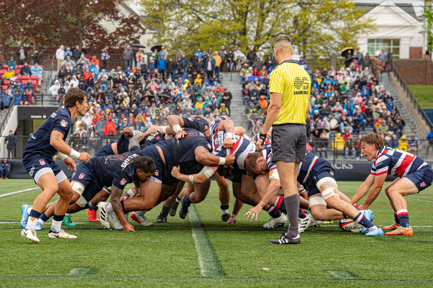 Rugby players forming a scrum during competitive match on green field with spectators watching.