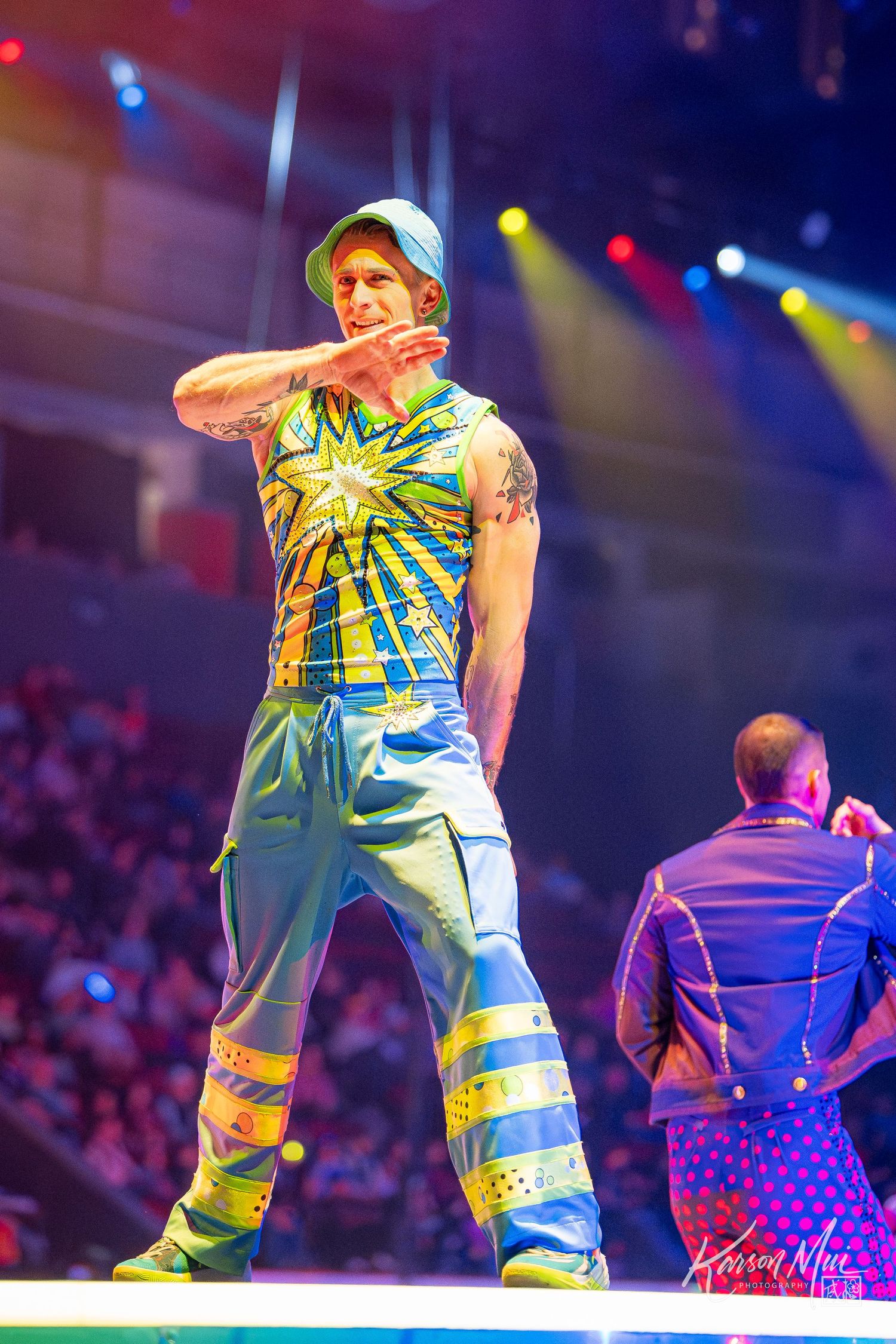 Male performer in colorful tie-dye costume dancing under stage lights at live show.