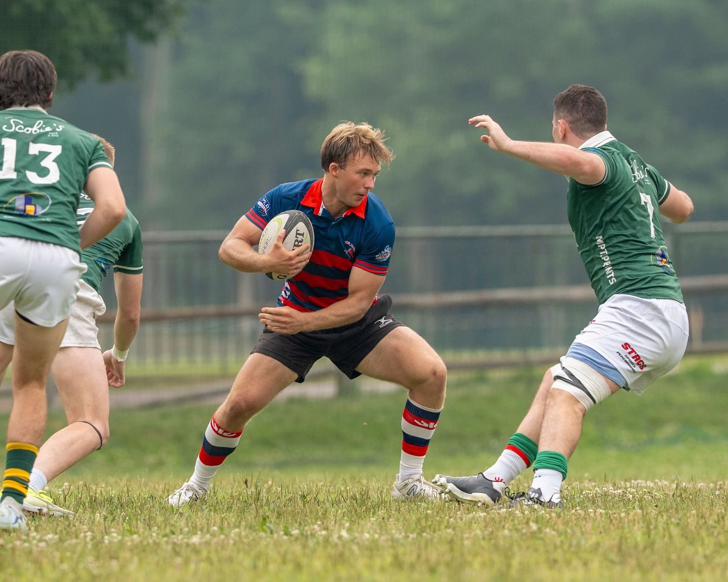 Rugby player in red and blue jersey carries ball while evading green-uniformed defenders on a grassy field.