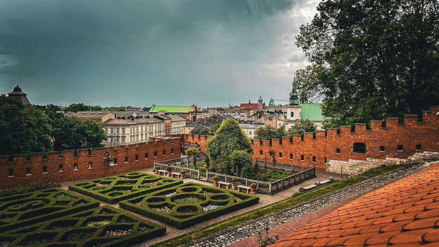 Aerial view of Wawel Castle's Renaissance garden with geometric hedges, red brick walls, and Krakow cityscape under stormy skies.Krakow, Pol