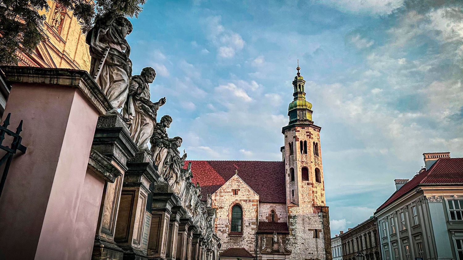 Historic European church with baroque statues, red-tiled roofs, and a green clock tower against a partly cloudy sky. Krakow, Poland