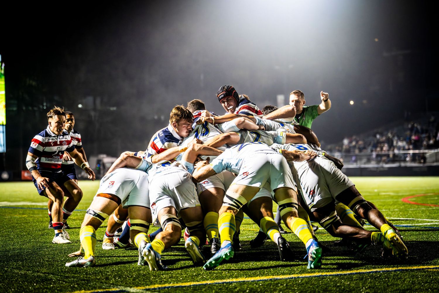 Rugby team forms a scrum under bright stadium lights during a night game.