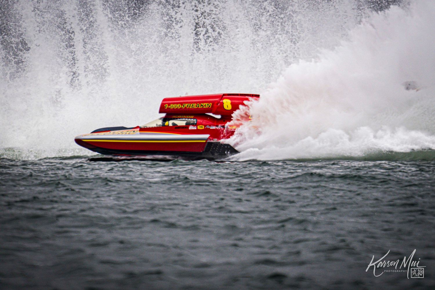 Red and gold hydroplane powerboat creates massive water spray while racing at high speed on open water.