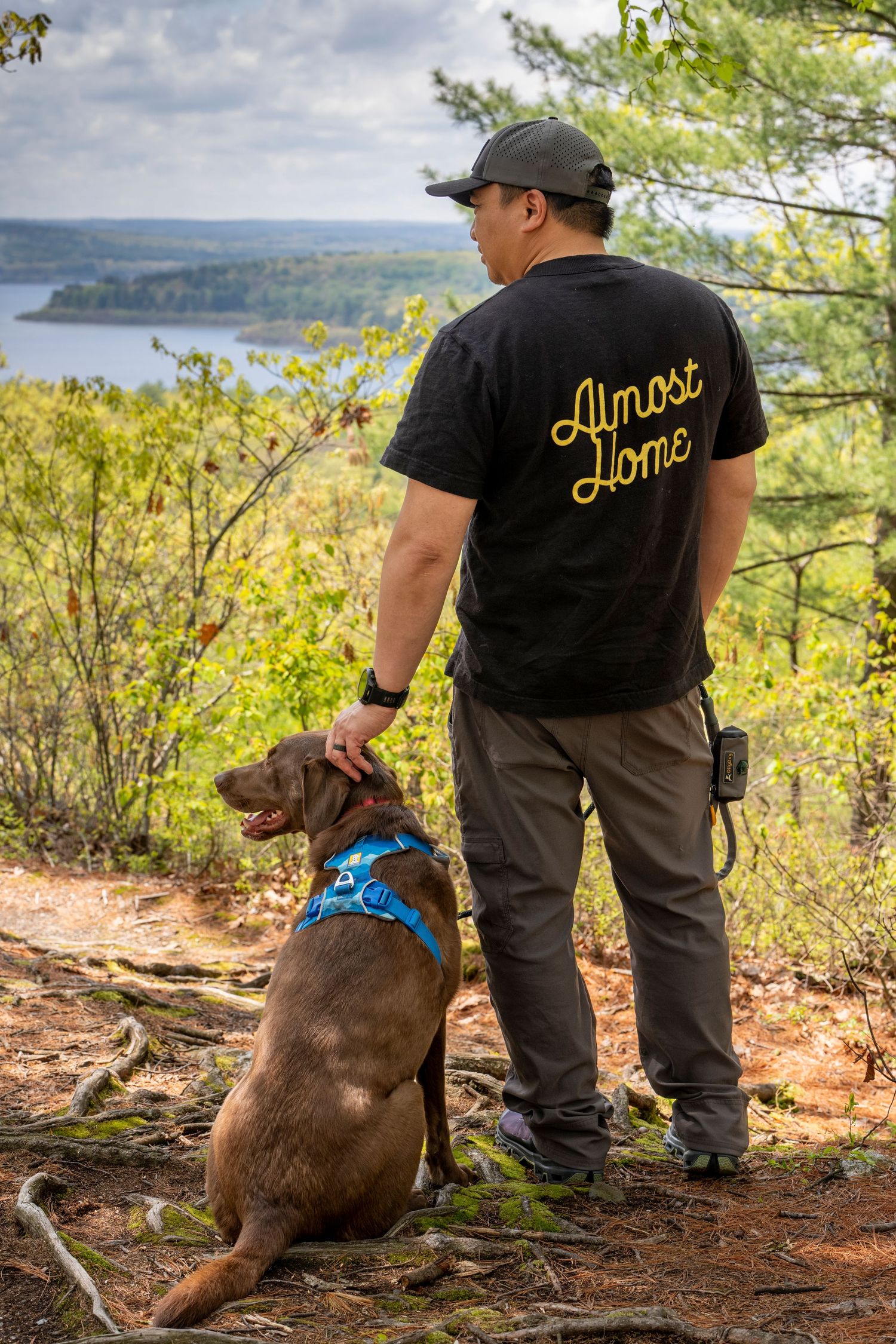 Man in Almost Home t-shirt stands with brown dog on scenic forest overlook above a lake.