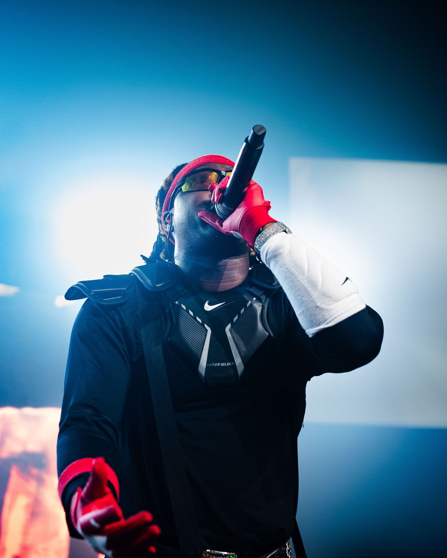 Performer in Nike gear and red accessories holds microphone on stage under dramatic blue lighting.