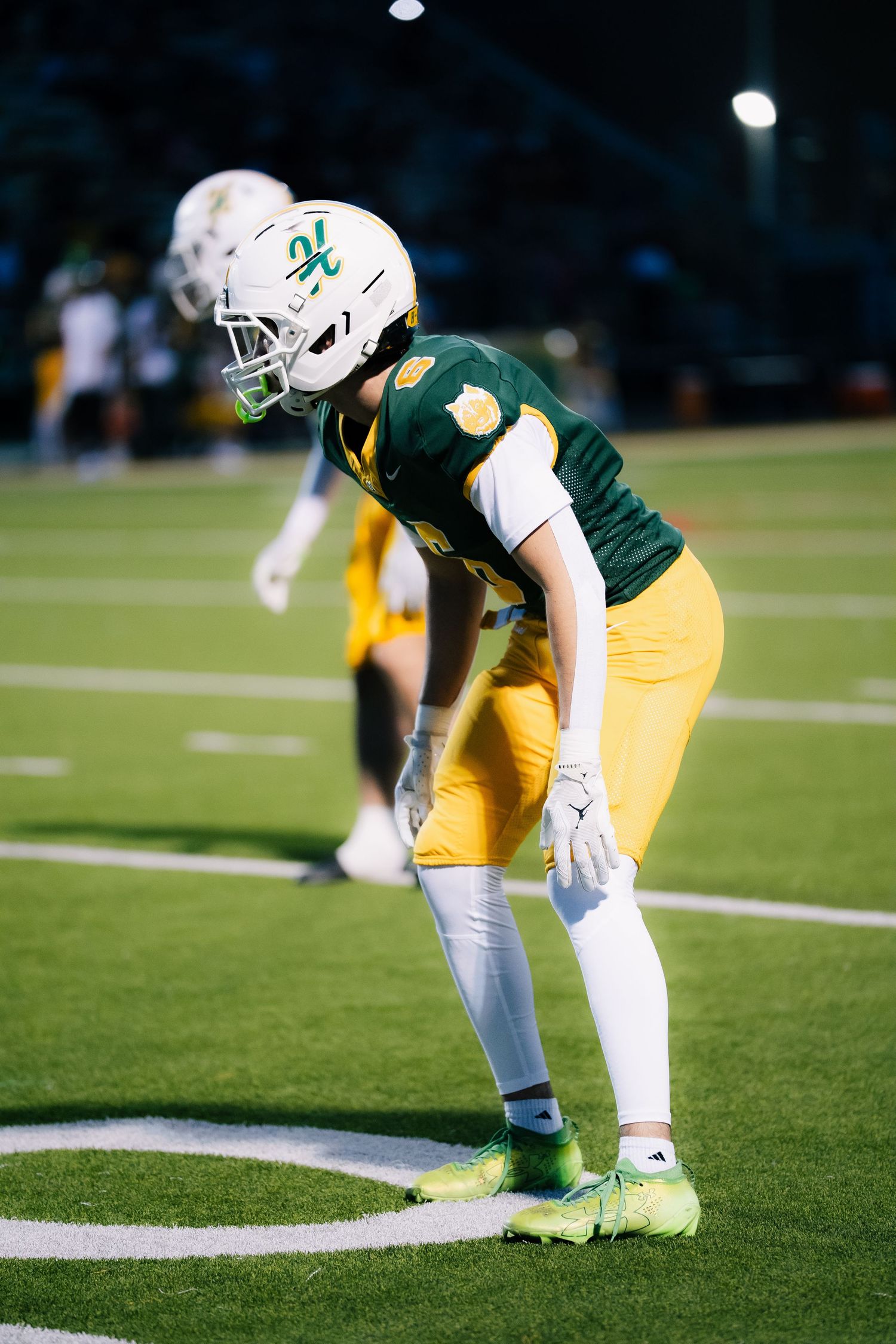 Football player in green and gold uniform crouches in ready position on field during a night game.
