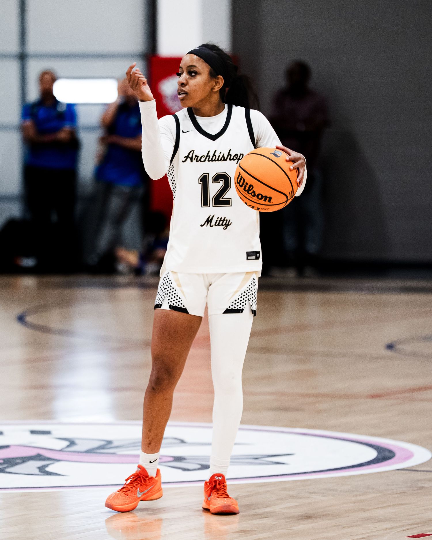 Female basketball player #12 in white Archbishop Mitty uniform dribbles Wilson basketball.