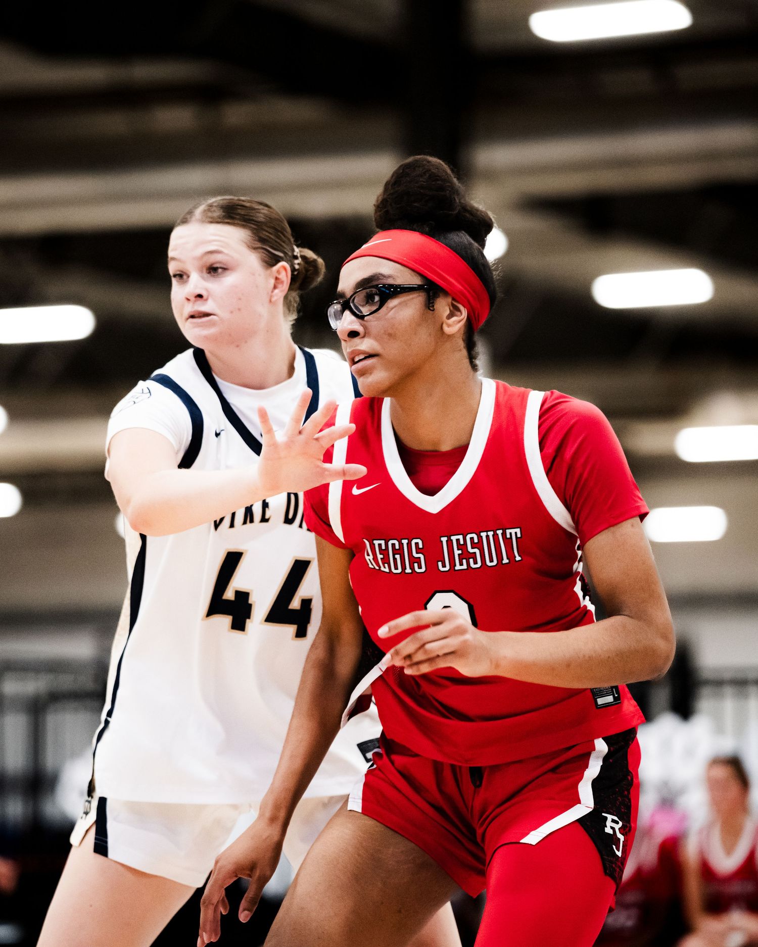 Two female basketball players compete, one in white #44 and one in red Regis Jesuit jersey, during an indoor game.