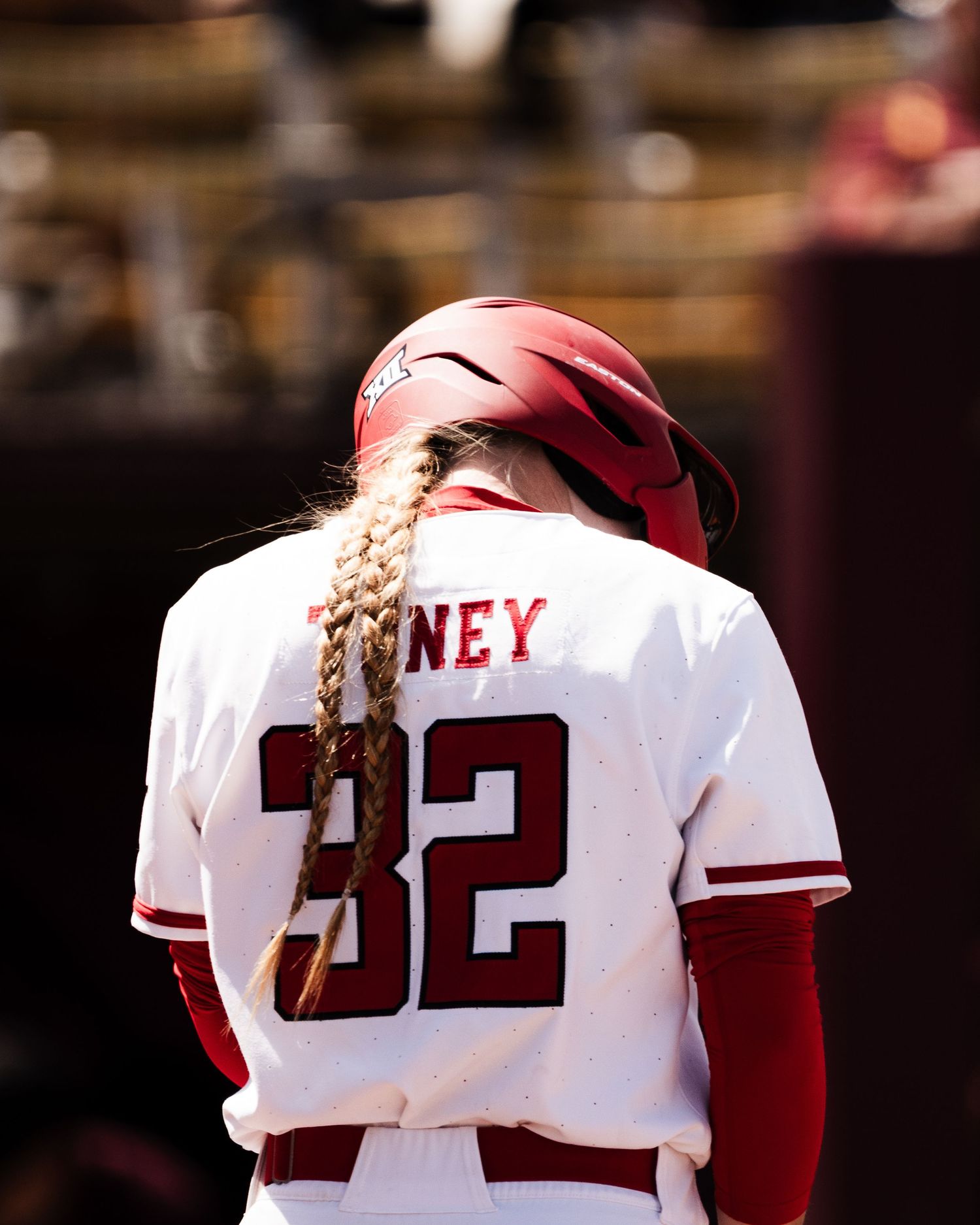 Softball player #32 ONEY in white uniform and red helmet with braided hair stands on field.