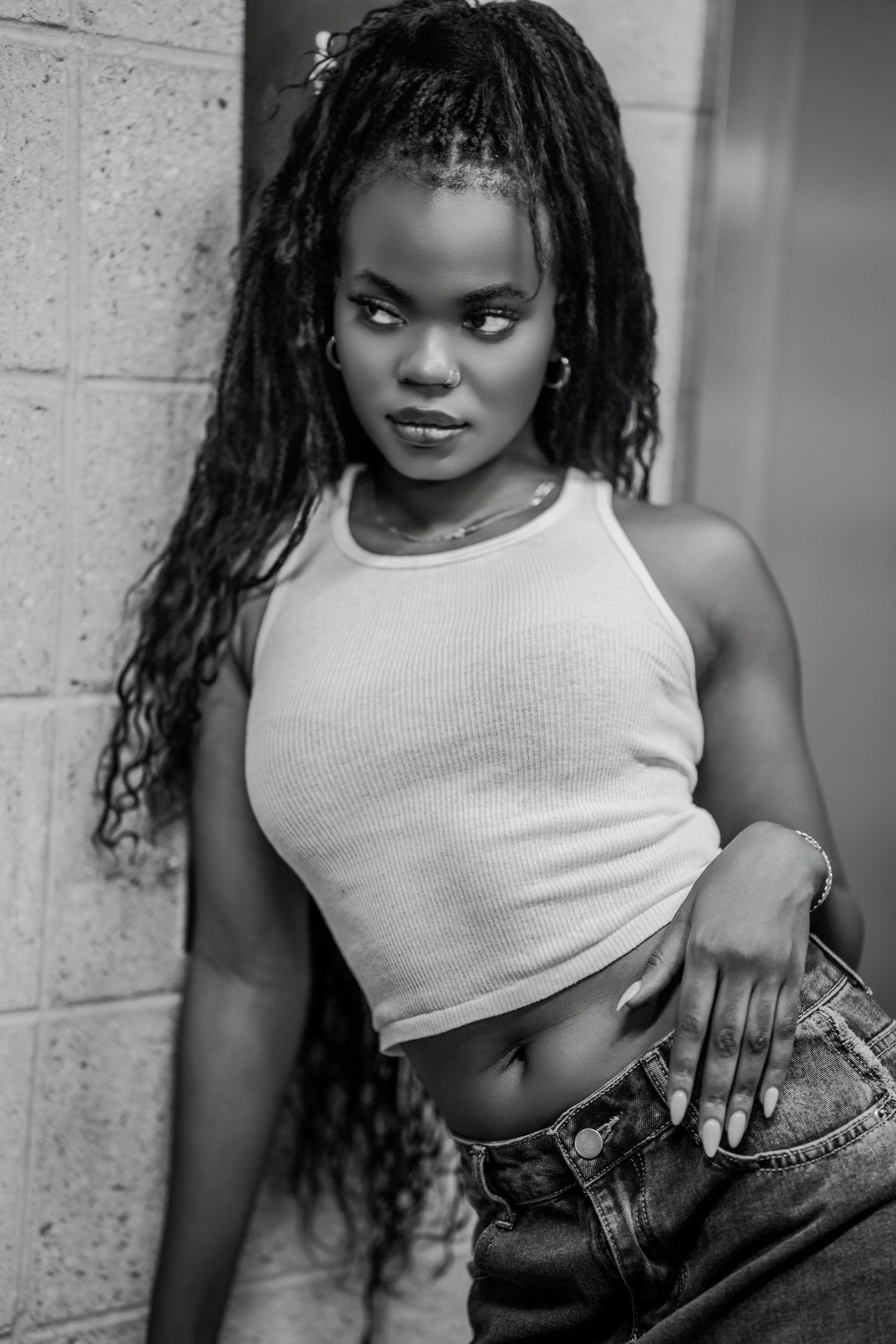 Black and white photo of a young woman with locs in a white crop top and jeans posing against a brick wall.