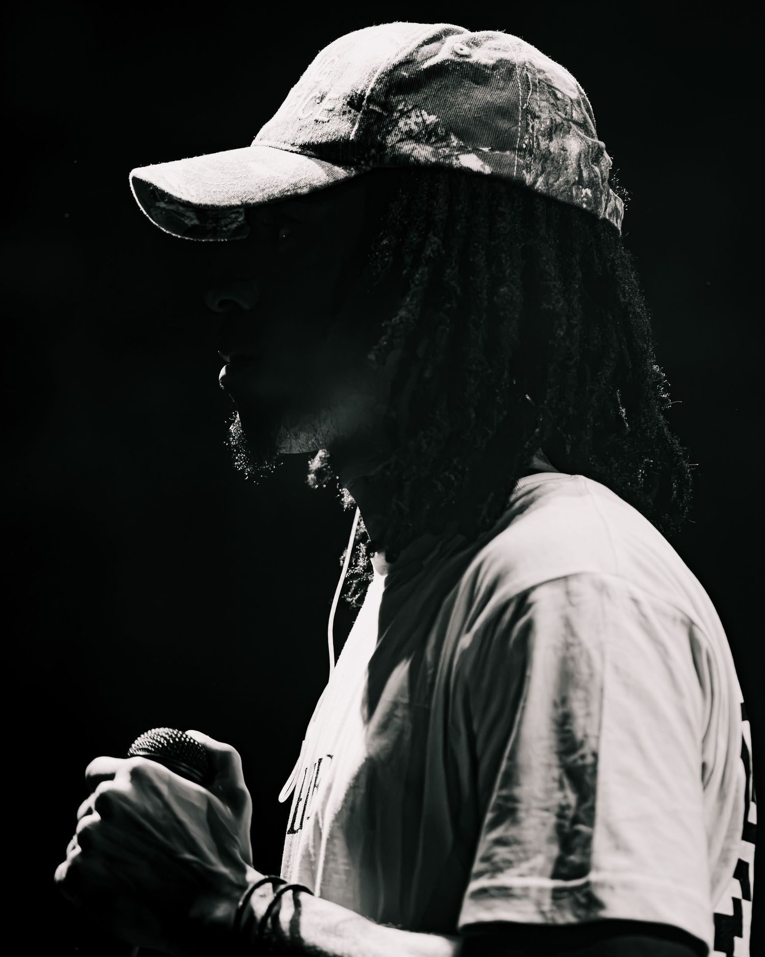 Black and white profile of a person with dreadlocks and a cap holding a microphone in dramatic stage lighting.