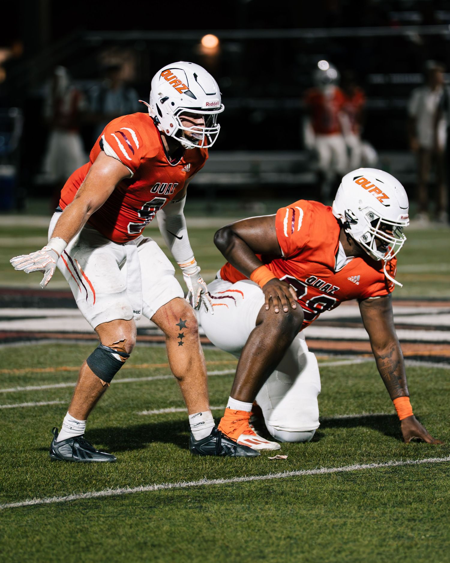Two football players in orange uniforms prepare at line of scrimmage during night game.