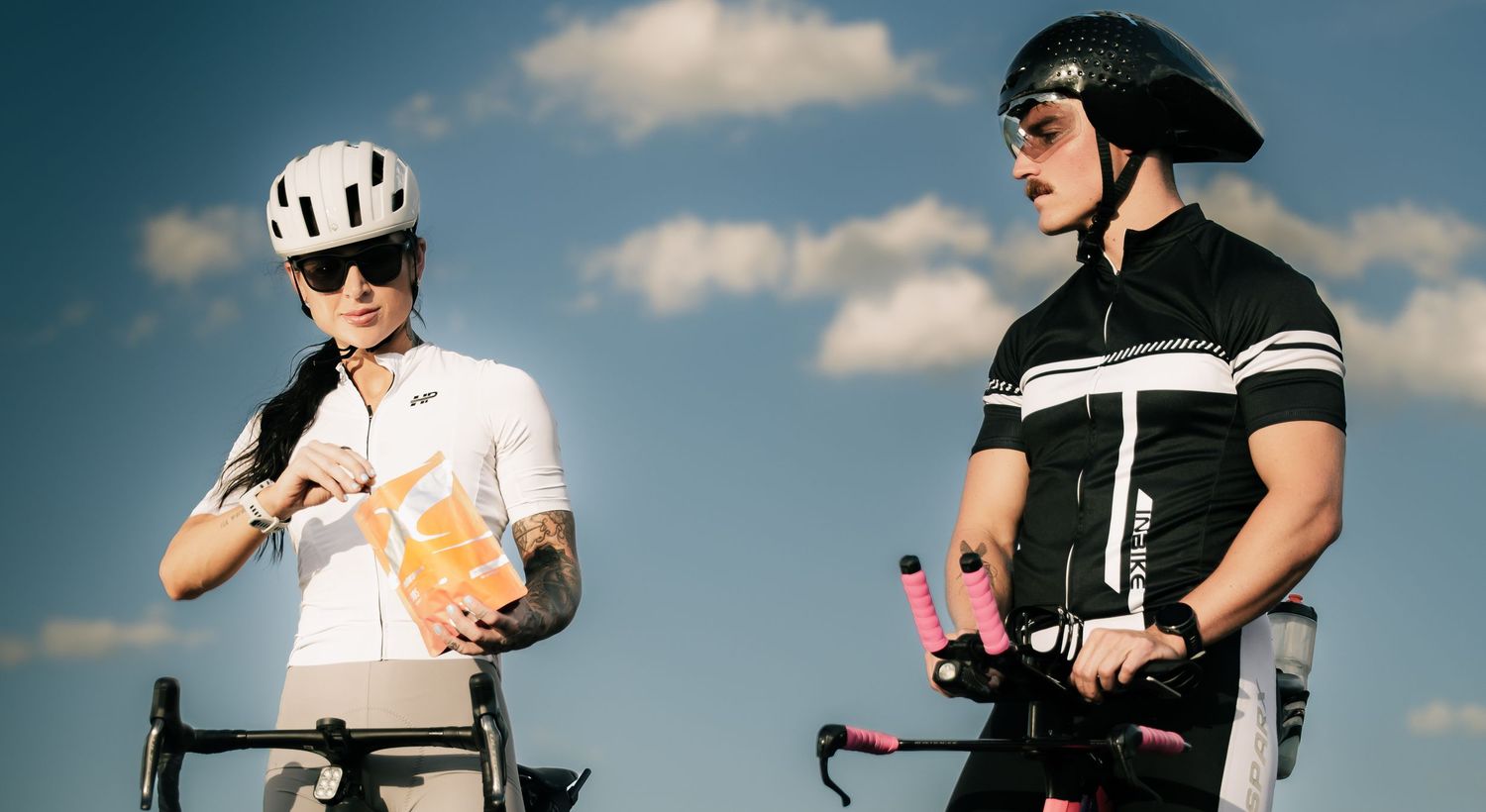 Two cyclists in helmets and sunglasses pause outdoors, one holding a snack, against a blue sky.
