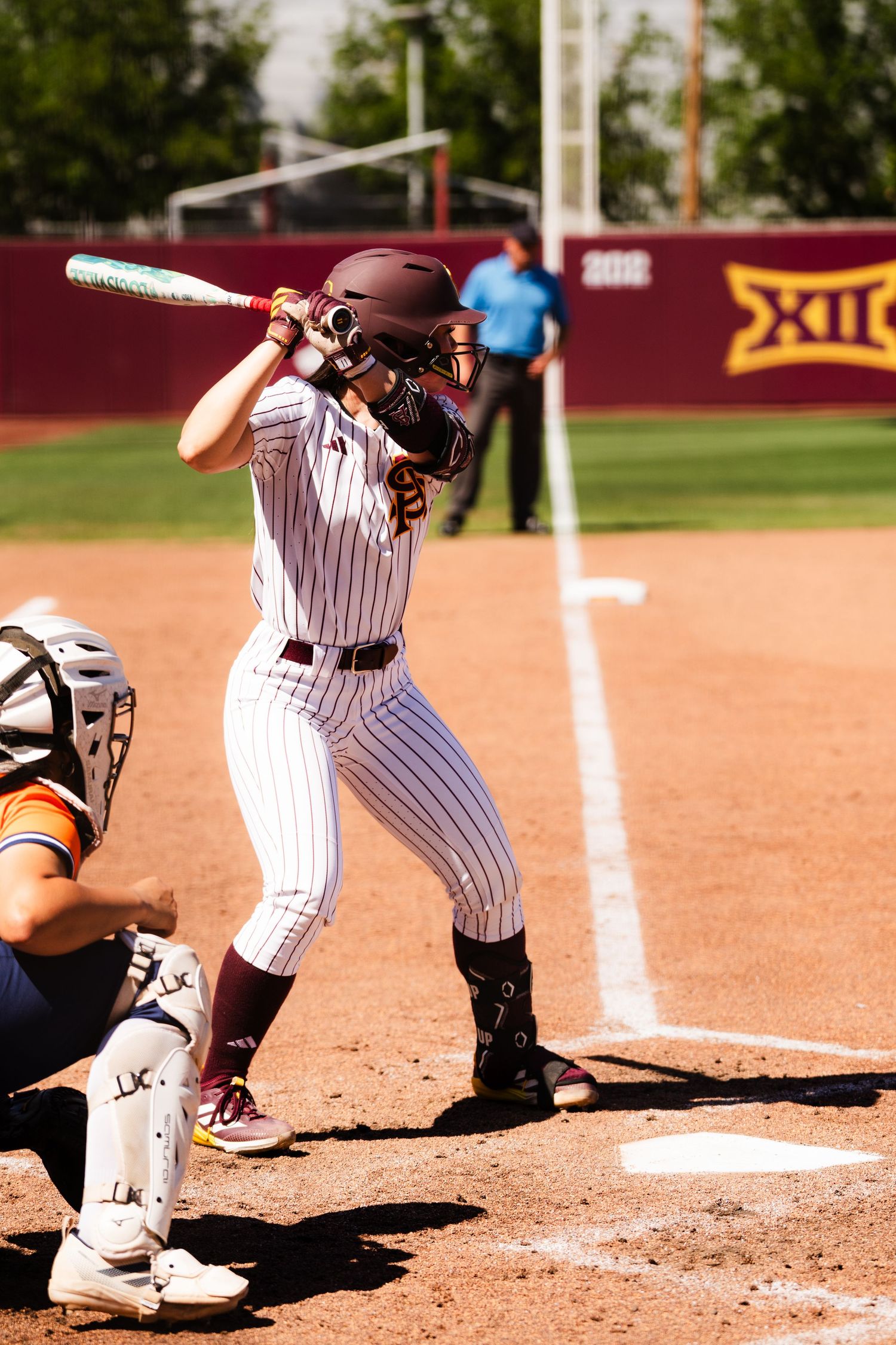 Female softball batter in maroon pinstripe uniform swings at pitch during a Big 12 college softball game.
