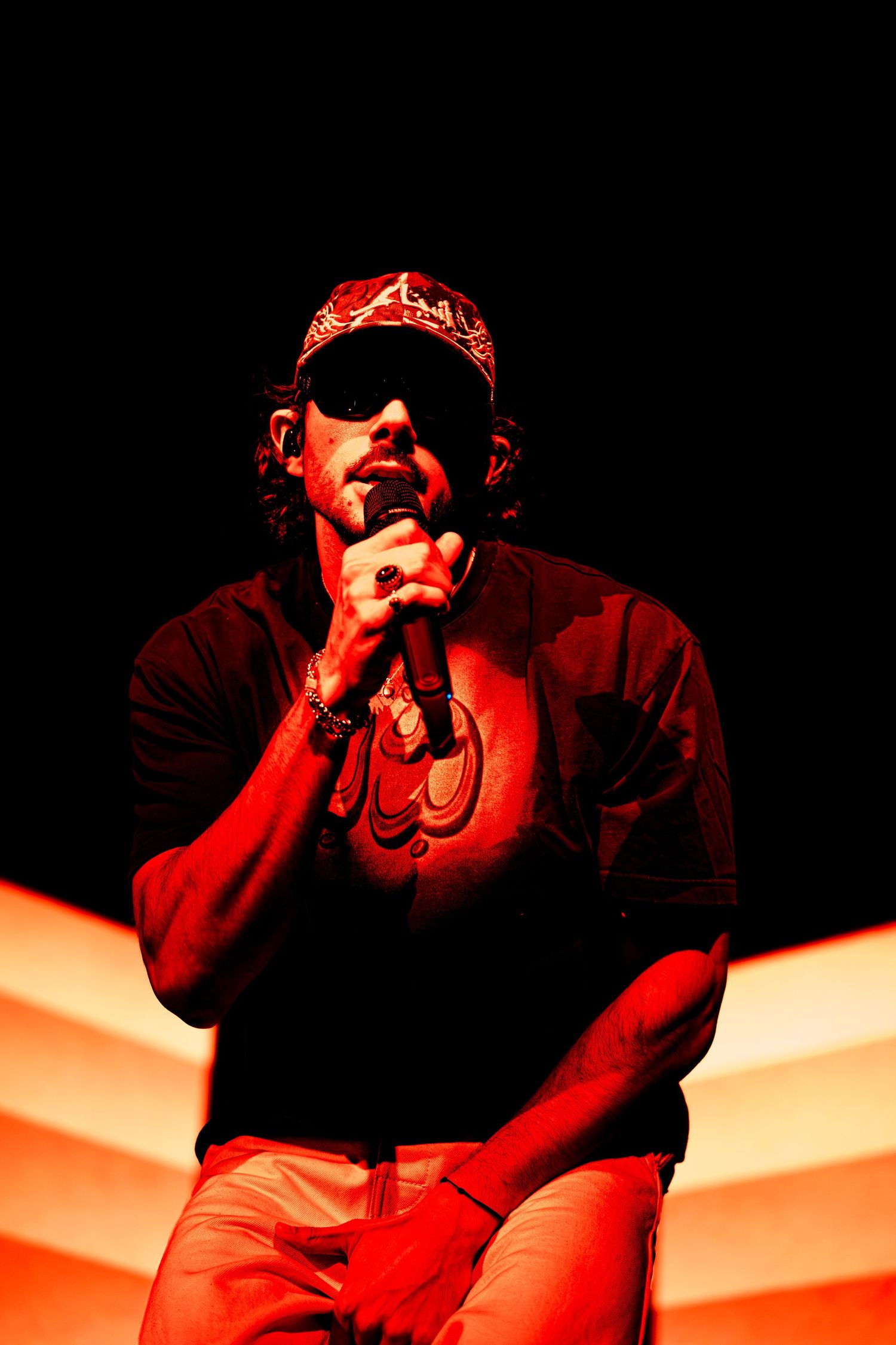 Performer in a bandana and sunglasses crouches on stage while singing into a microphone, bathed in dramatic red and orange concert lighting.