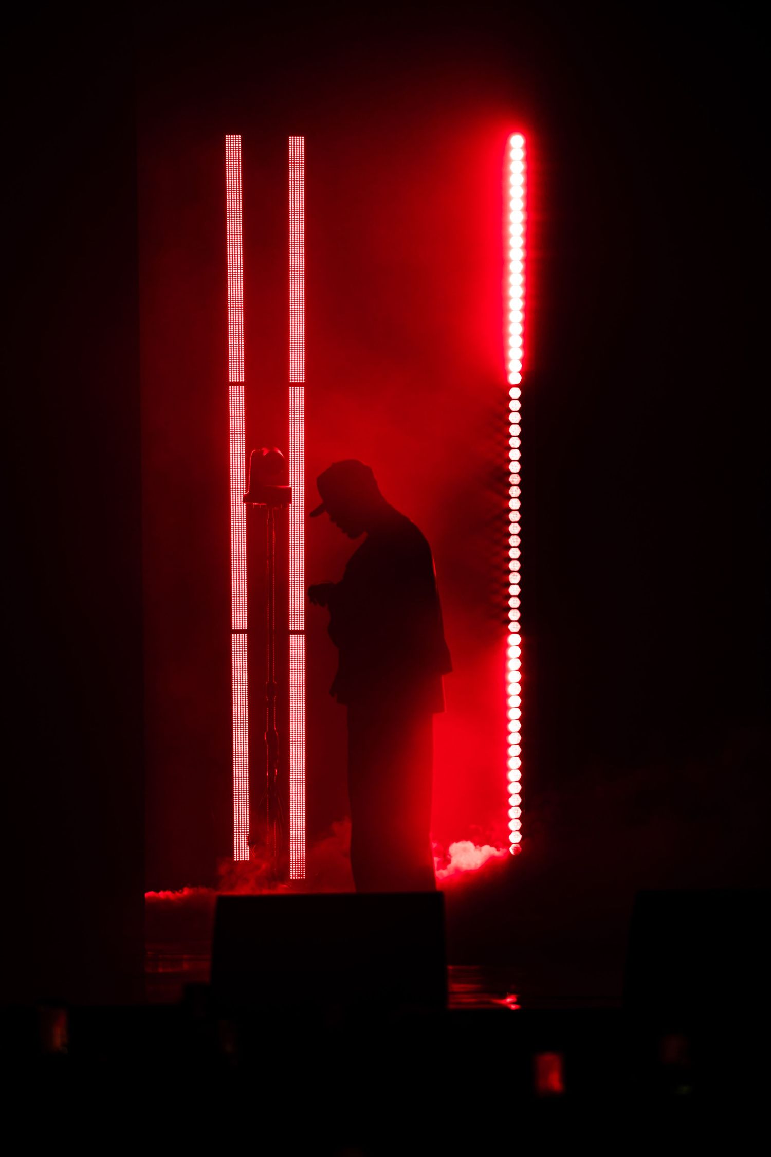 Silhouette of a performer on a dark stage surrounded by haze, glowing red vertical LED light bars and white light strips behind them.