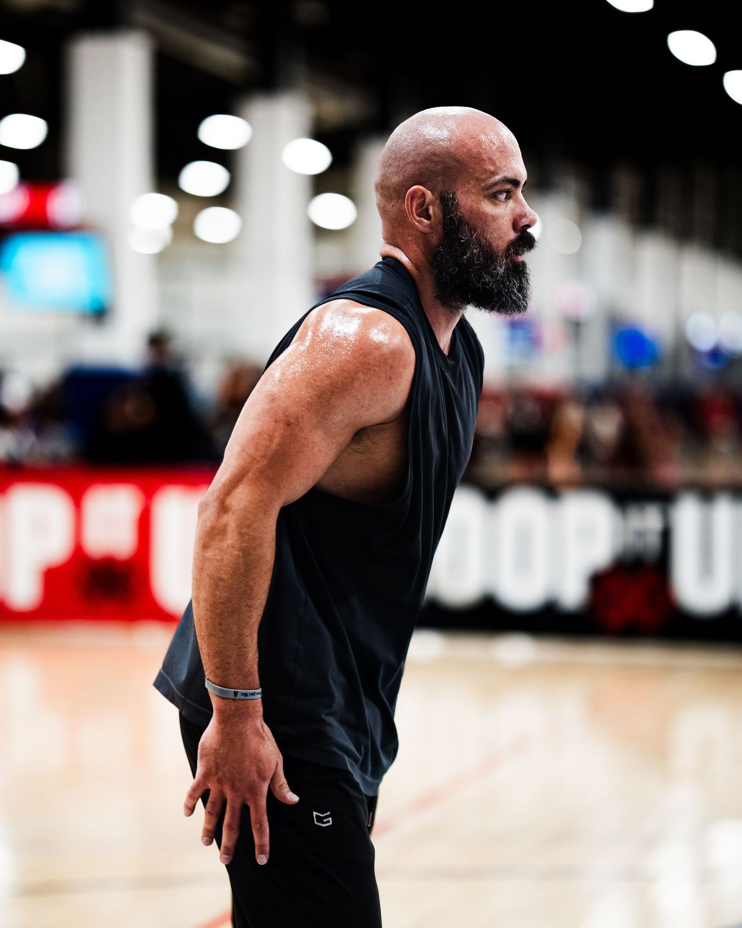 Muscular bald man with beard wearing black sleeveless shirt stands on basketball court at indoor sports event.